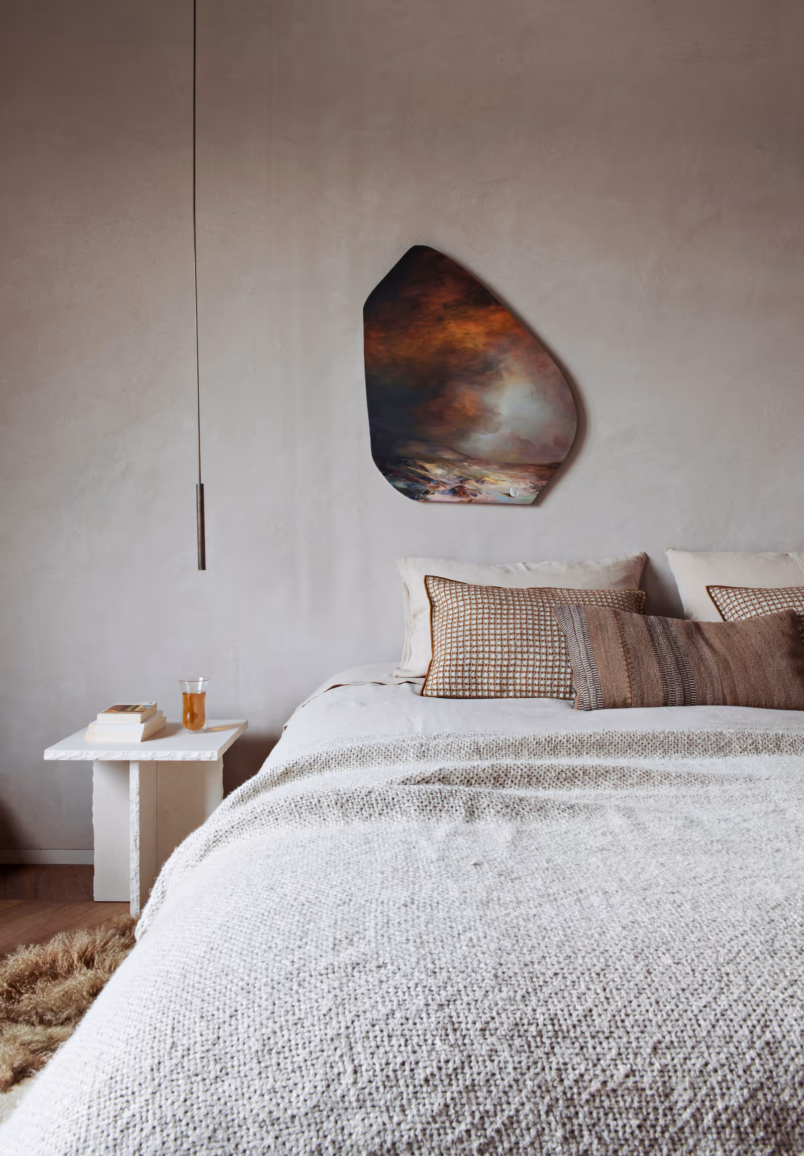 Chambre minimaliste avec une couverture beige texturée, des coussins à motifs, des œuvres d'art abstraites au mur et une table d'appoint blanche sur laquelle sont posés des livres et un verre de thé.