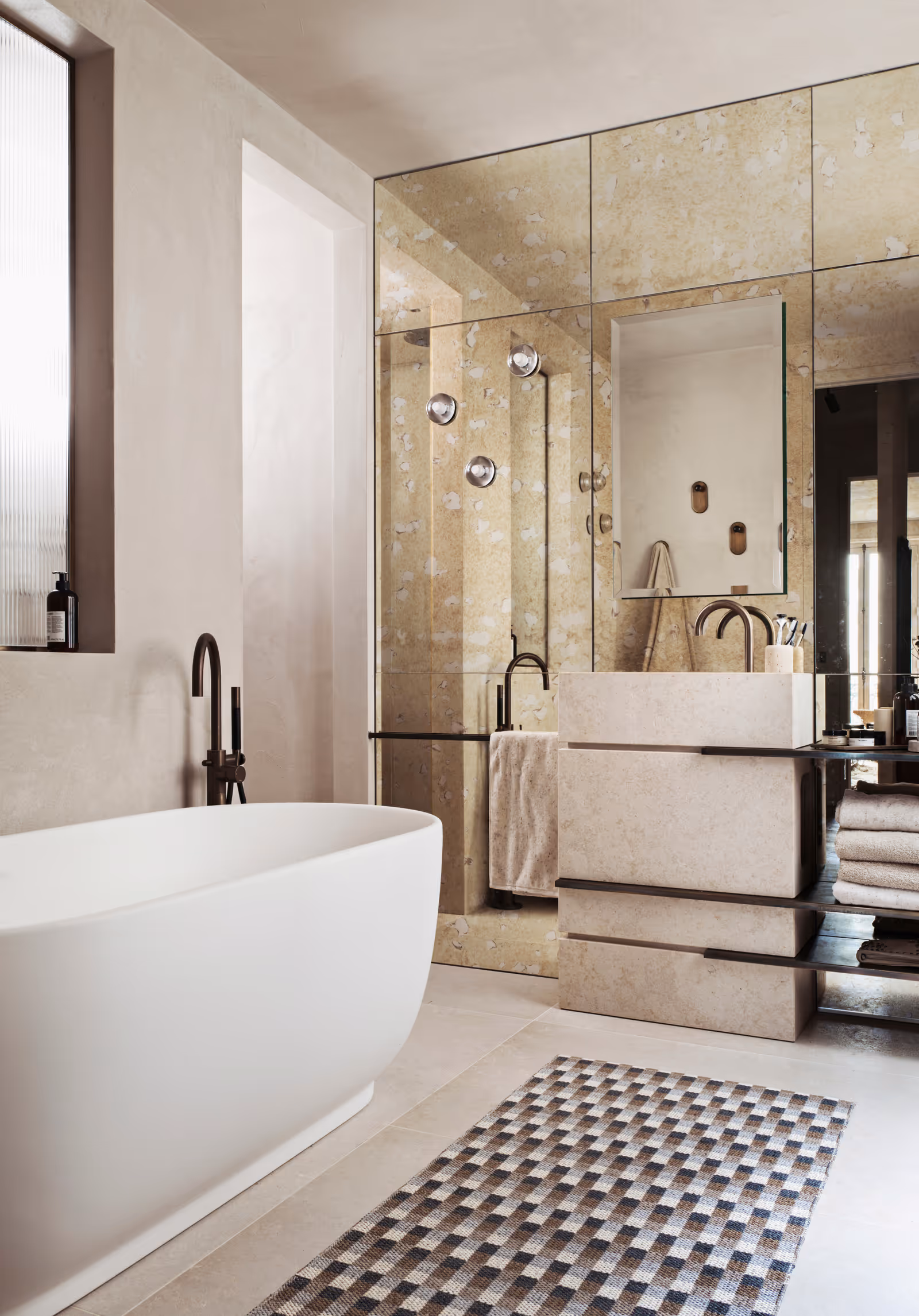 Salle de bain moderne avec baignoire îlot blanche, meuble-lavabo en pierre beige, mur recouvert de miroirs et tapis à carreaux.