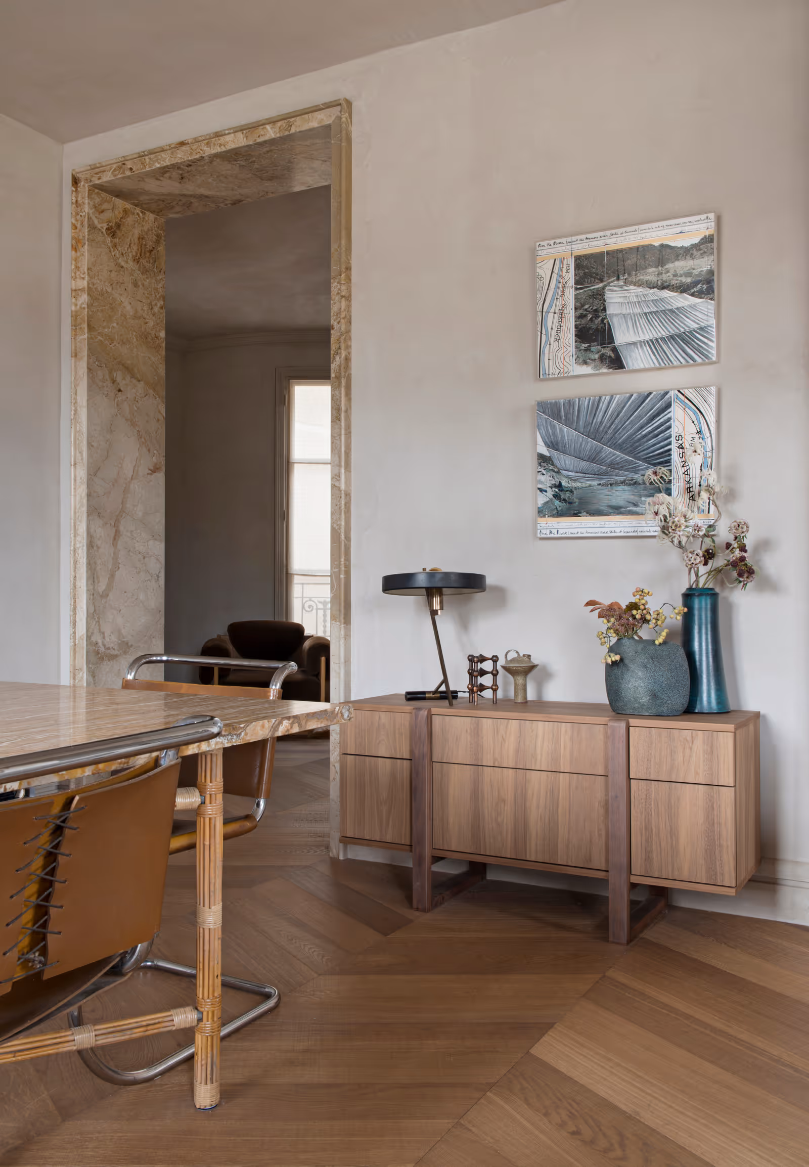 Coin salle à manger moderne avec une table en bois et des chaises en cuir marron, un buffet en bois avec des vases décoratifs et une lampe, et deux peintures abstraites représentant des paysages sur un mur beige.