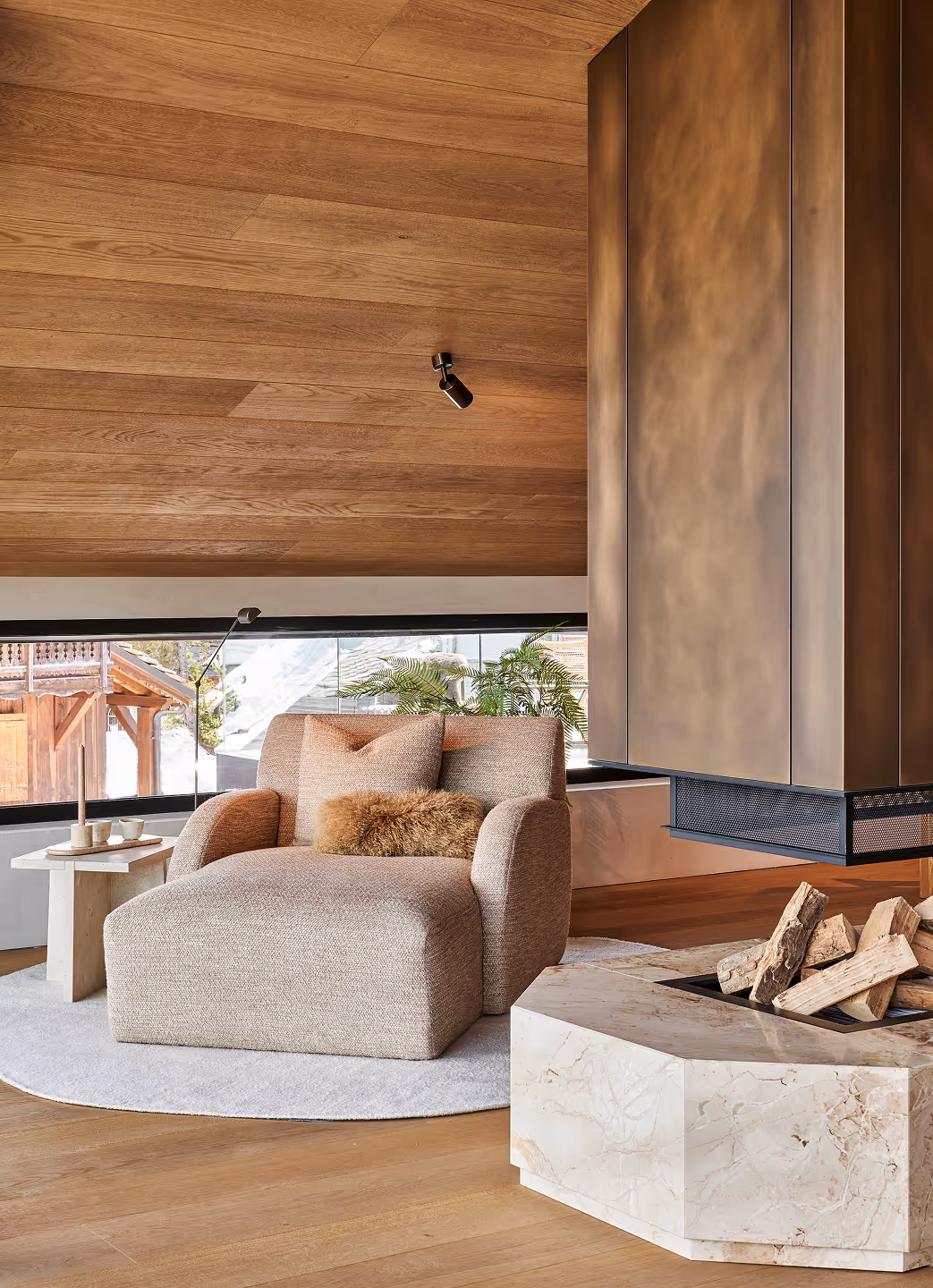 Fauteuil beige confortable avec coussins sur un tapis rond près d'une cheminée moderne avec du bois empilé dans une pièce lambrissée.