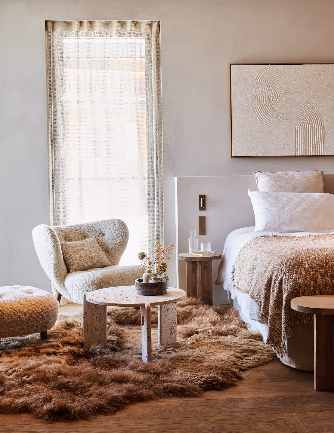 Coin chambre confortable avec un fauteuil beige texturé, une table basse ronde en céramique posée sur un tapis brun moelleux, et un lit recouvert de draps blancs et d'un plaid brun.