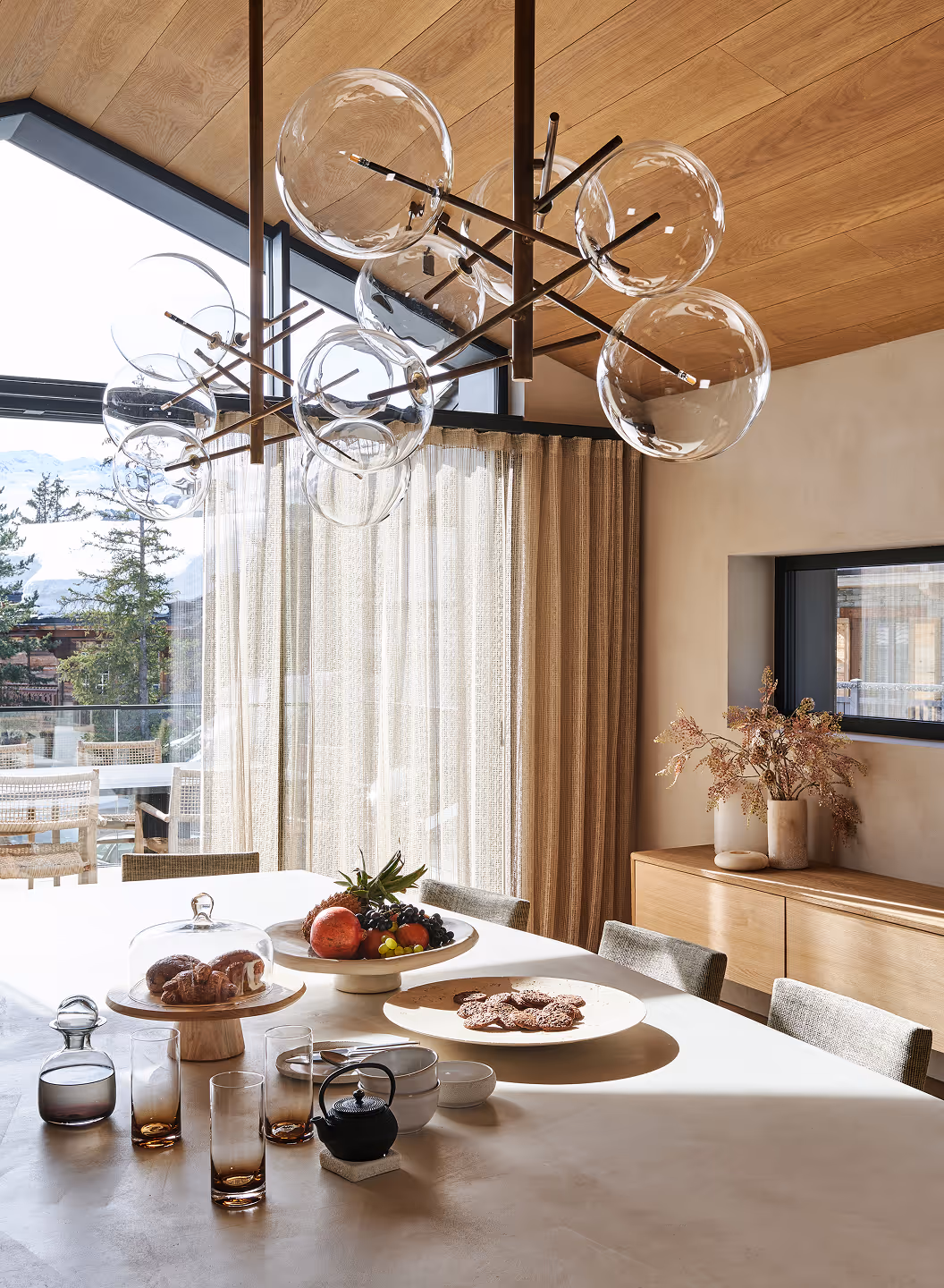 Salle à manger moderne avec une table blanche dressée avec des fruits, des pâtisseries sous verre, une théière et des verres, agrémentée de grands luminaires suspendus en forme de globe en verre et de baies vitrées ornées de rideaux transparents.