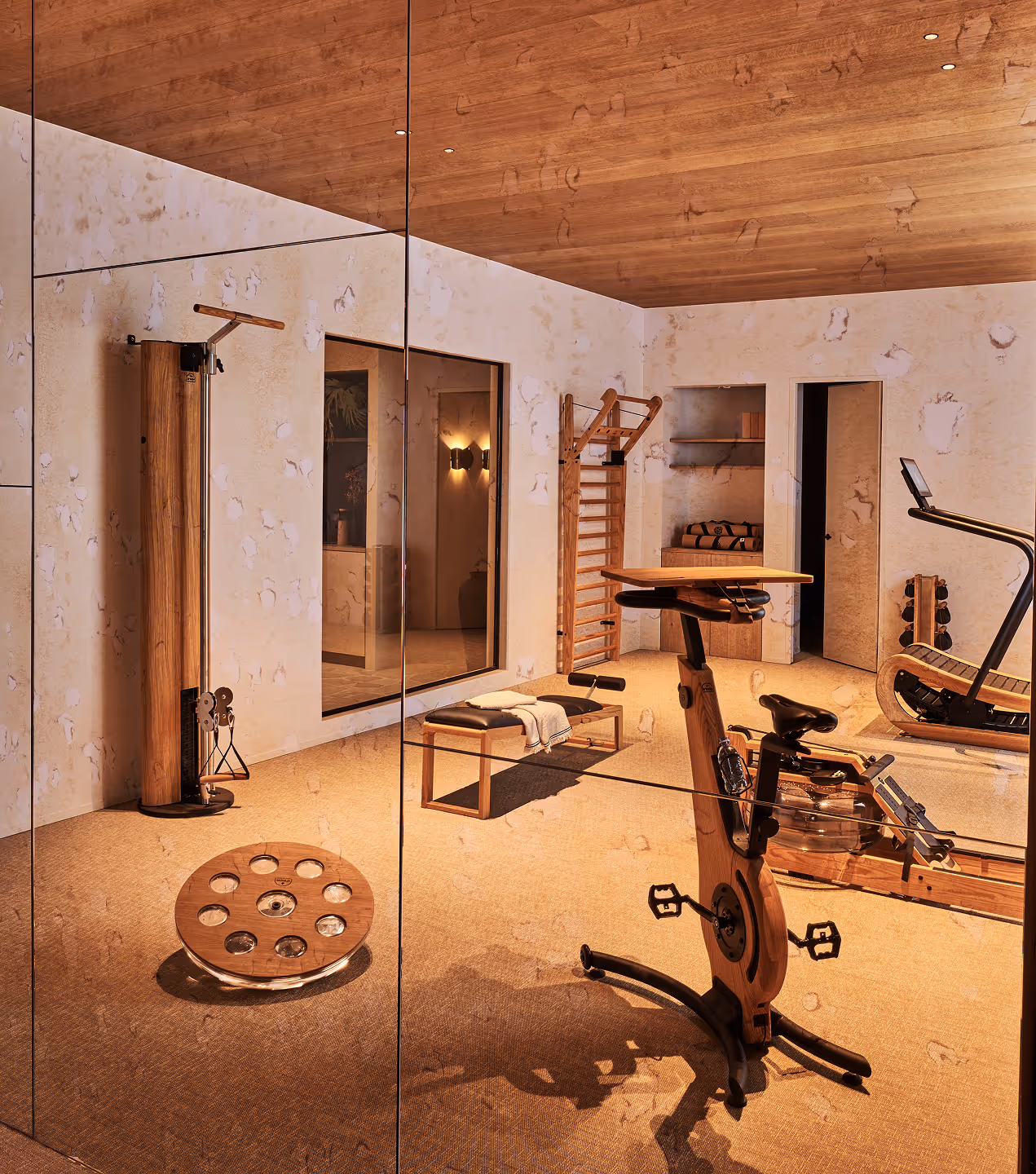 Salle de sport moderne à domicile avec vélo d'appartement en bois, banc avec serviette, poulie et mur recouvert de miroirs reflétant l'équipement.