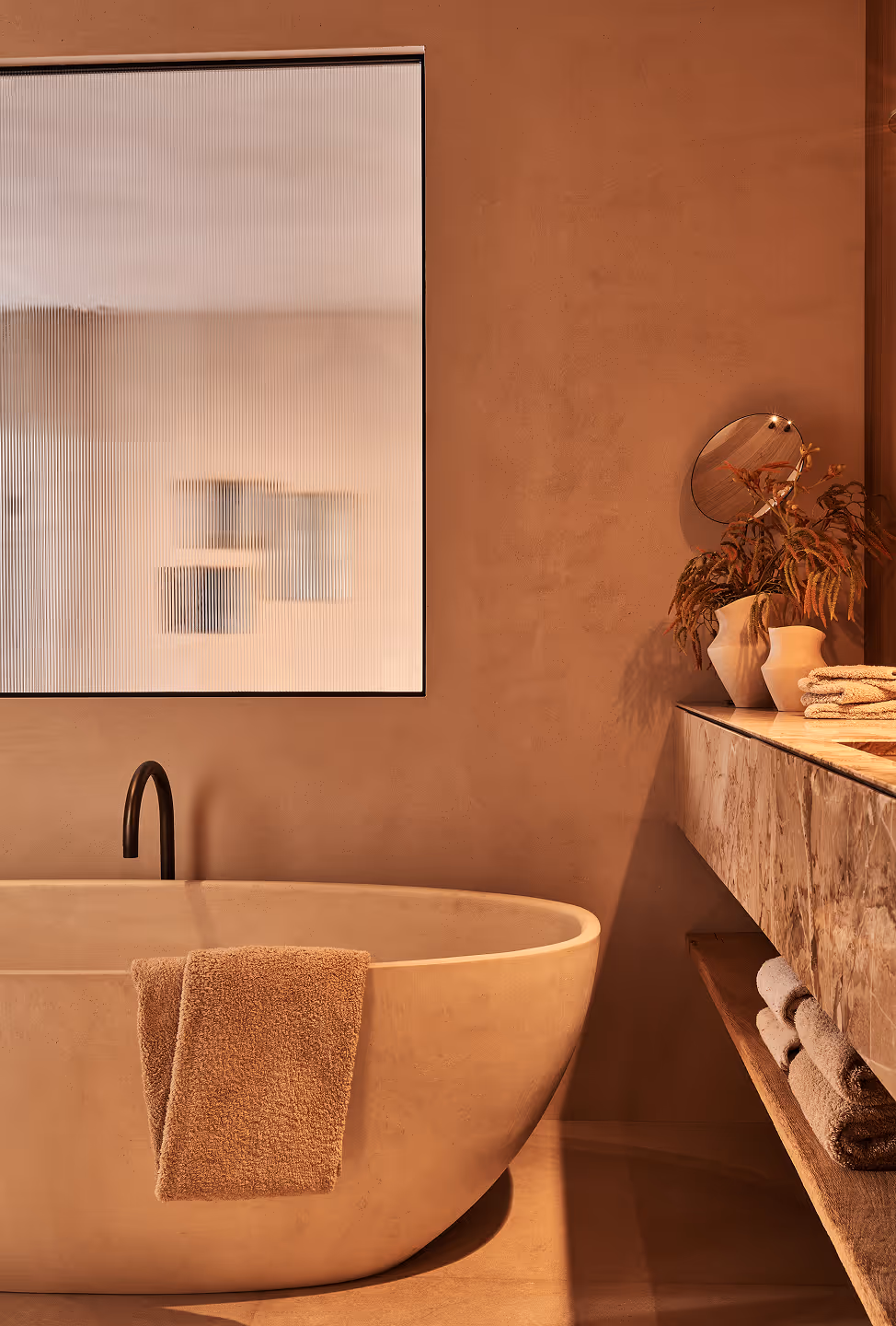 Salle de bain moderne avec une baignoire îlot beige, une serviette marron posée dessus, un robinet noir et un plan de travail en marbre avec des serviettes pliées et des vases beiges contenant des plantes séchées.
