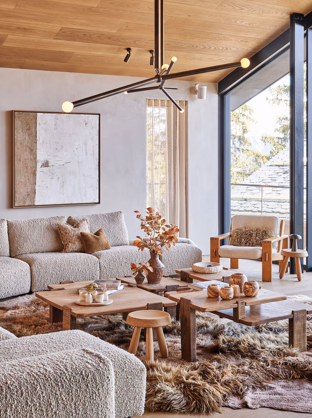 Salon confortable avec canapé d'angle beige texturé, tables basses en bois, tapis en fourrure, suspension moderne et grande fenêtre donnant sur les arbres à l'extérieur.