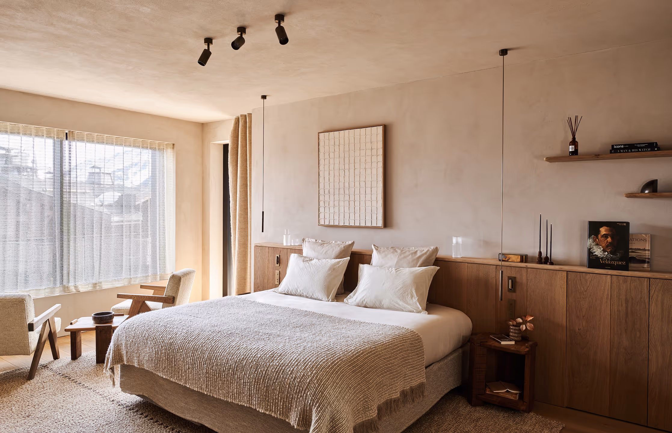 Chambre moderne avec un grand lit recouvert de couvertures beiges, deux fauteuils près d'une grande fenêtre avec des rideaux transparents et des armoires encastrées en bois le long du mur.