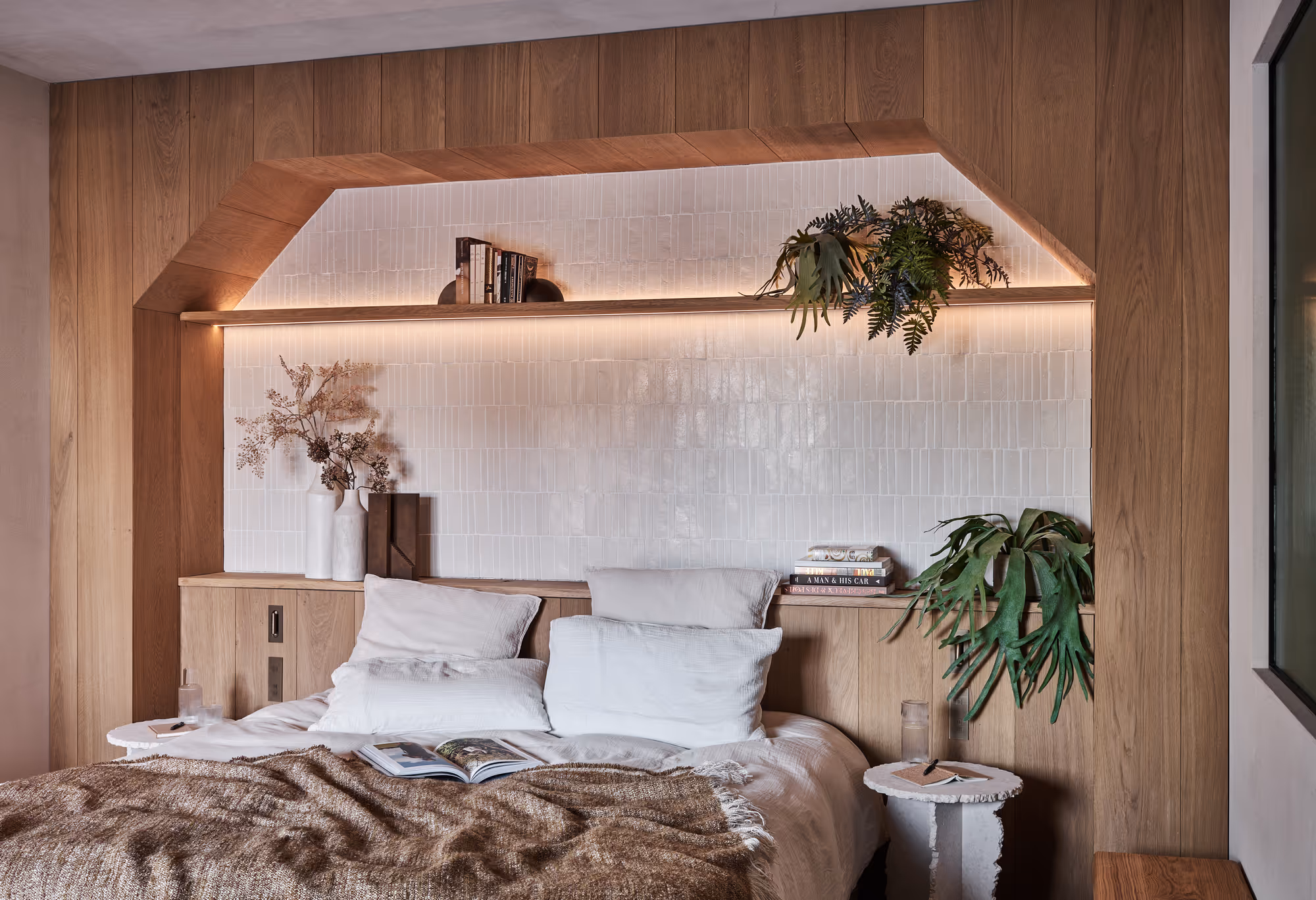 Chambre moderne et confortable avec un mur d'accent carrelé blanc encadré de bois, un lit avec des oreillers blancs, une couverture beige, des plantes, des livres et des tables de chevet.