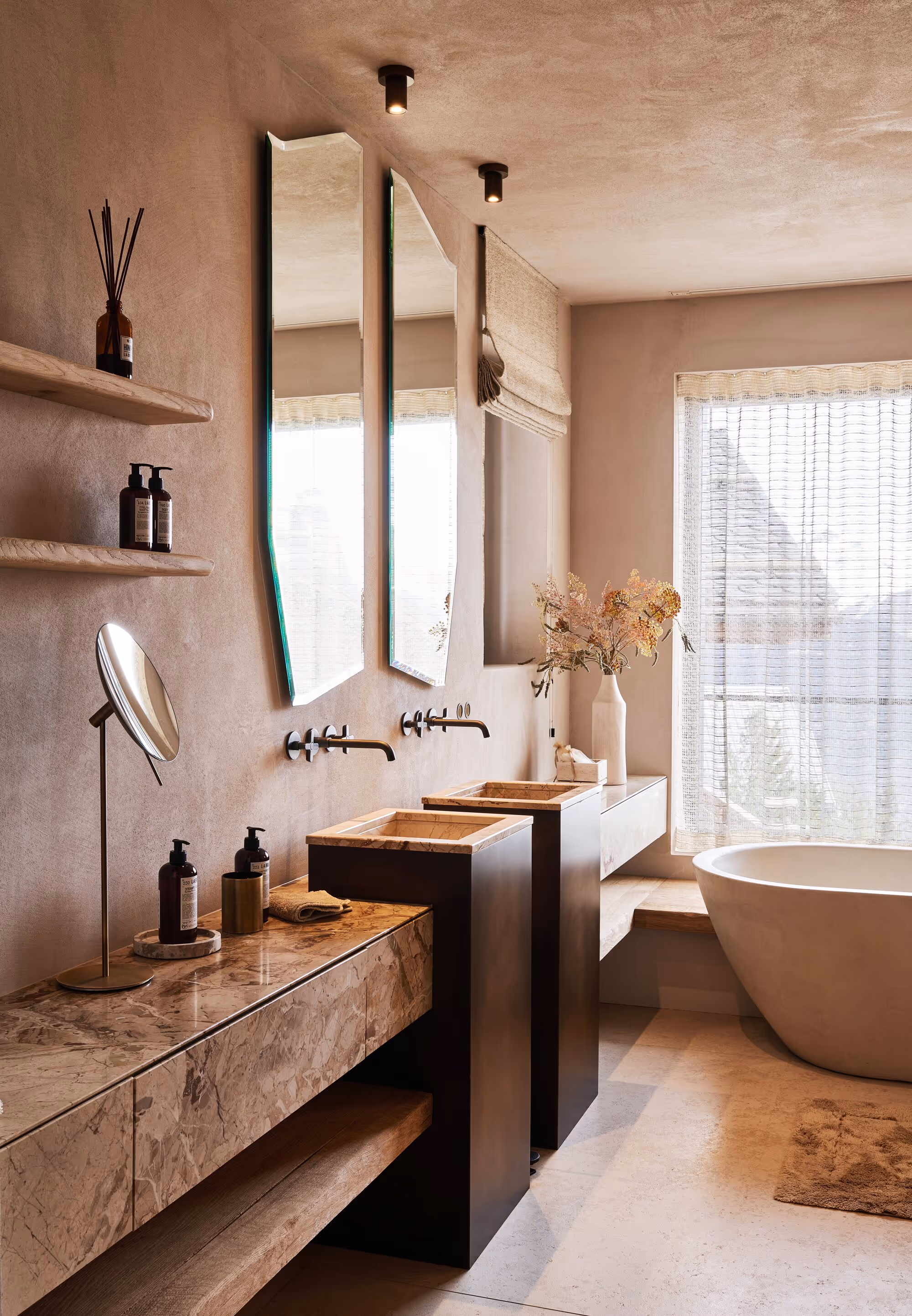 Salle de bain moderne avec deux lavabos rectangulaires en marbre sur des socles sombres, deux miroirs muraux asymétriques, une baignoire îlot près d'une grande fenêtre avec des rideaux transparents et une décoration dans les tons beiges.