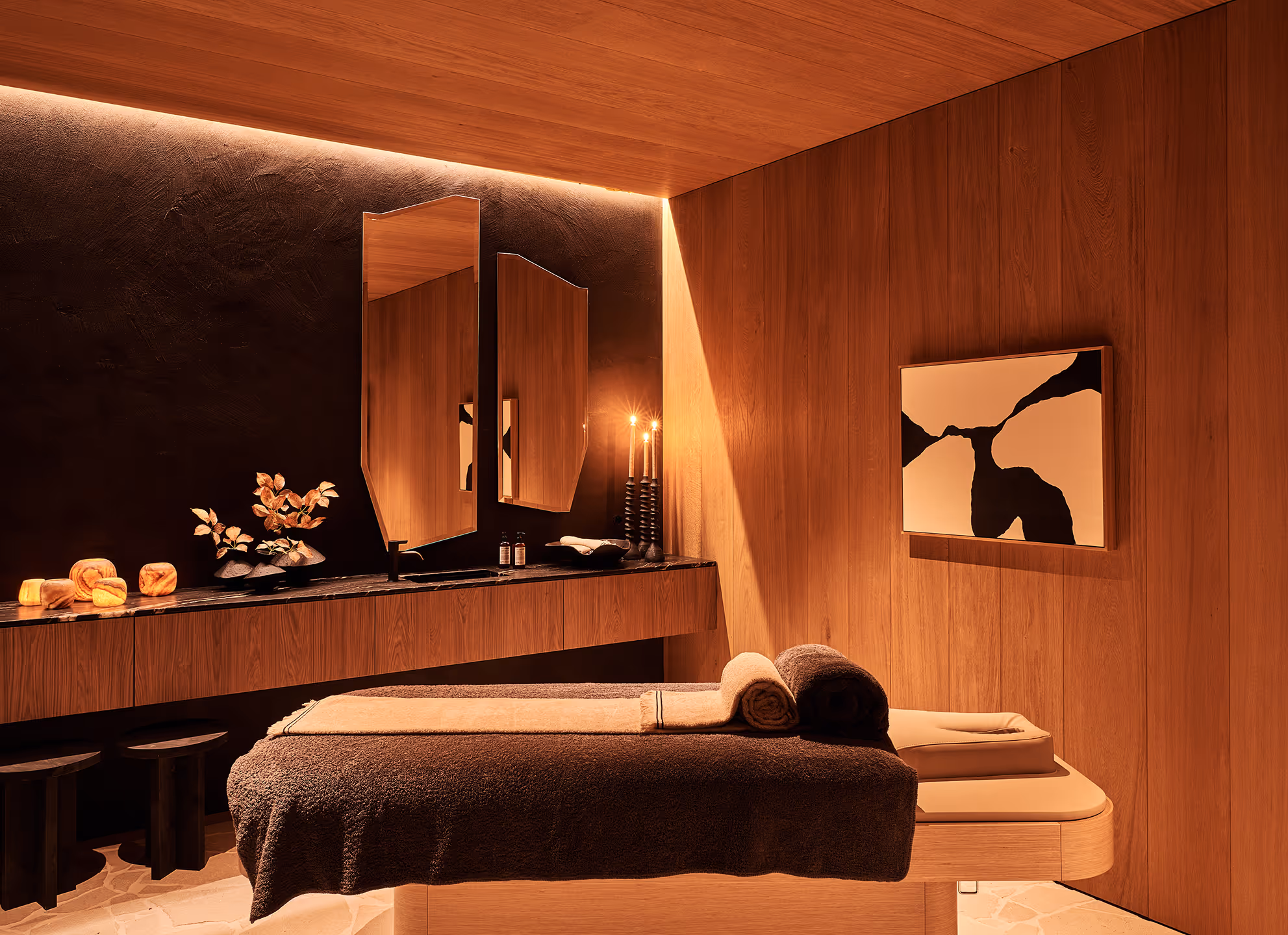 Salle de massage confortable avec des murs en bois, une table de massage rembourrée recouverte de serviettes marron, des œuvres d'art abstraites aux murs et un éclairage d'ambiance tamisé.