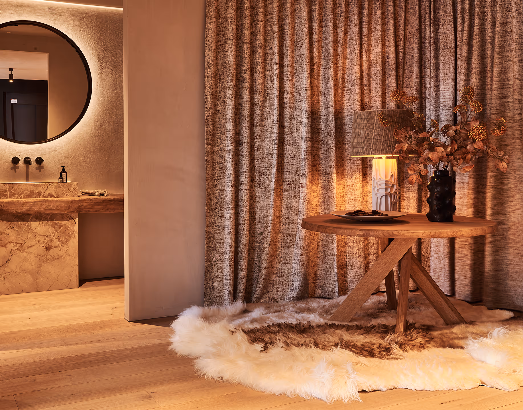 Coin confortable avec une table ronde en bois sur laquelle sont posés un vase noir contenant des fleurs séchées et une lampe allumée, le tout posé sur un tapis rond moelleux, à côté de rideaux beiges texturés et avec une vue partielle sur une salle de bain en marbre avec un miroir rond.
