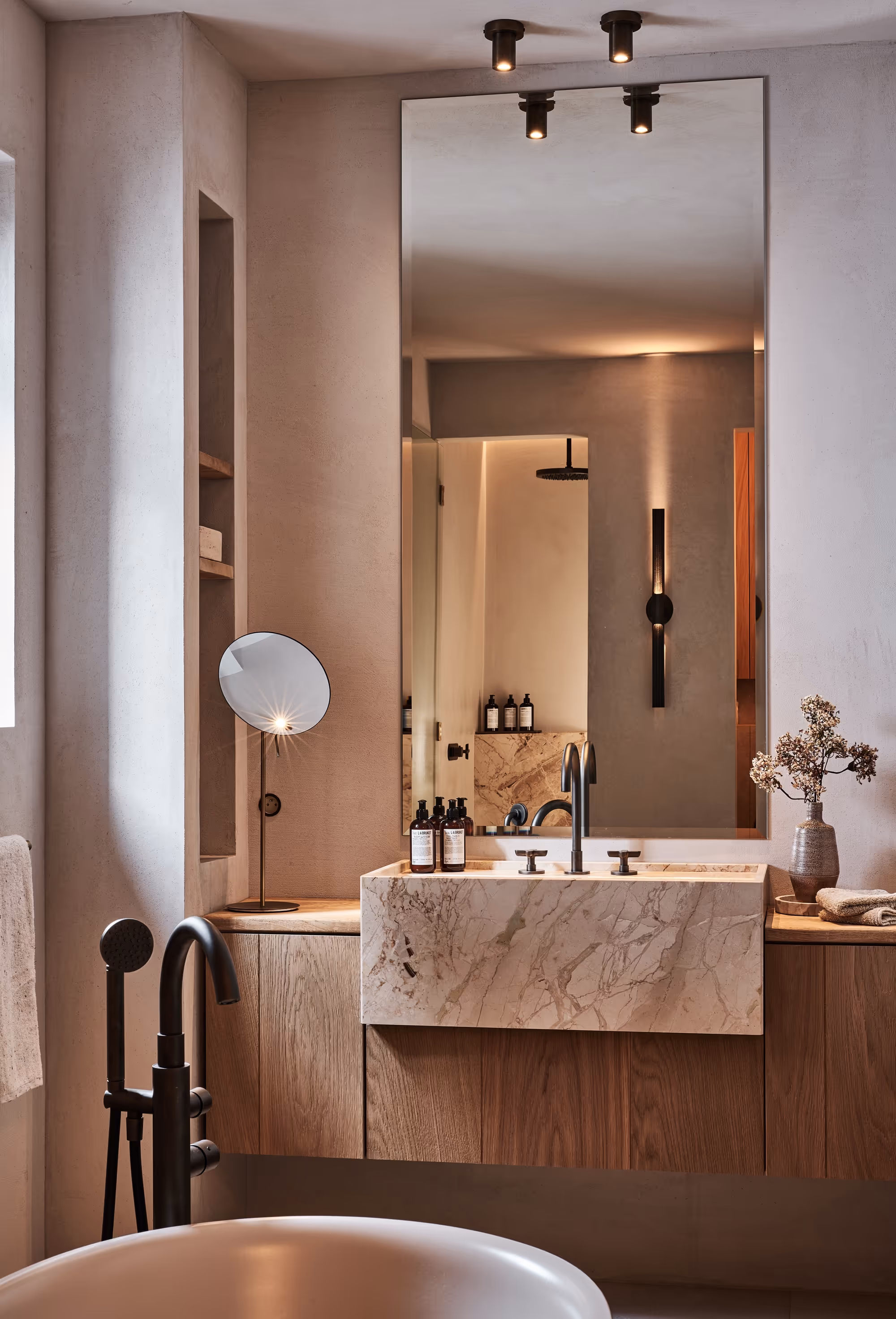 Meuble-lavabo moderne avec lavabo en marbre, grand miroir, accessoires noirs mats, miroir rond sur pied et vase décoratif avec fleurs séchées.