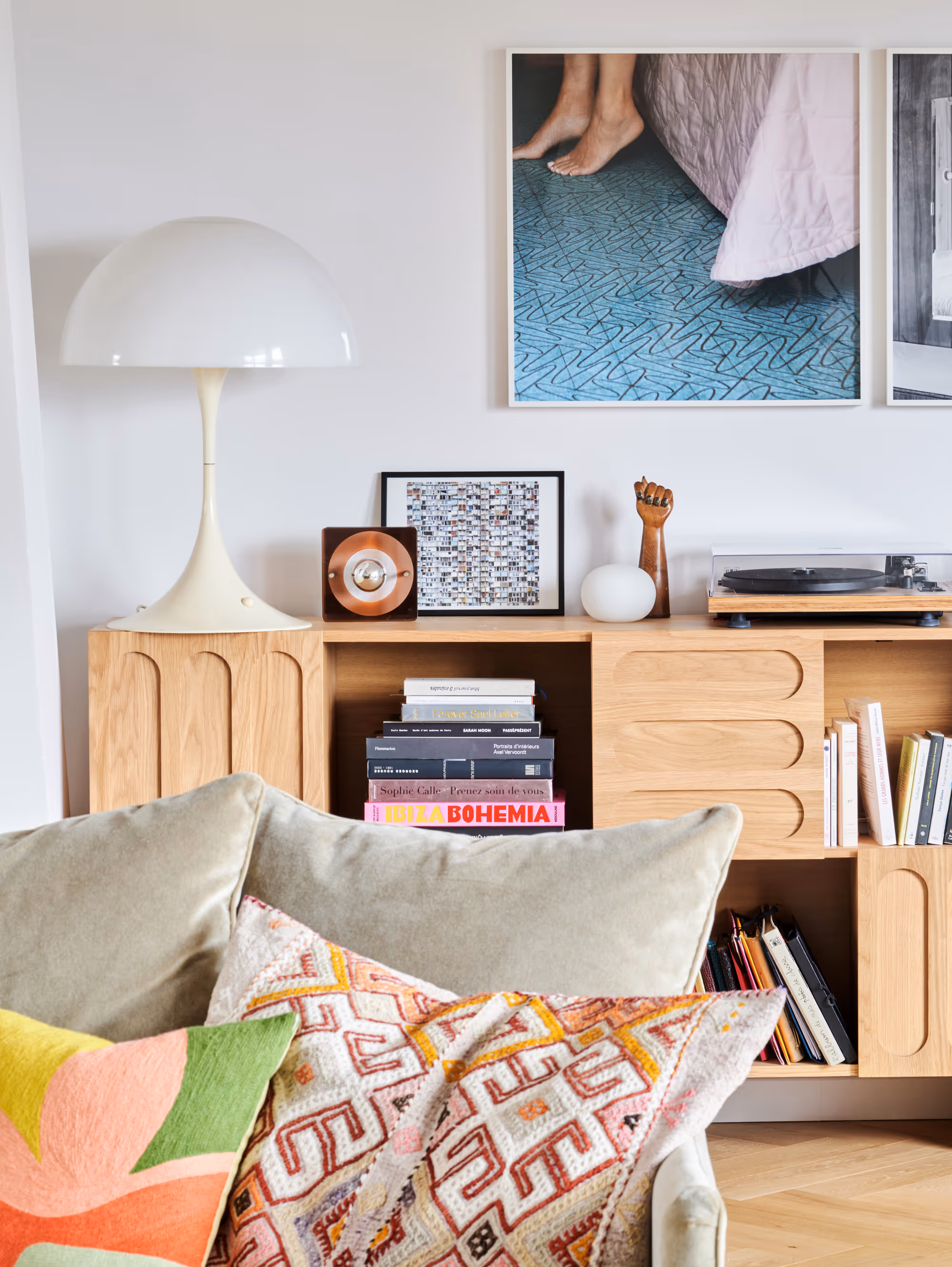 Salon moderne d'un duplex à Paris avec des étagères en bois clair contenant des livres, des œuvres d'art, une platine vinyle et des objets décoratifs derrière un canapé orné de coussins aux motifs colorés.