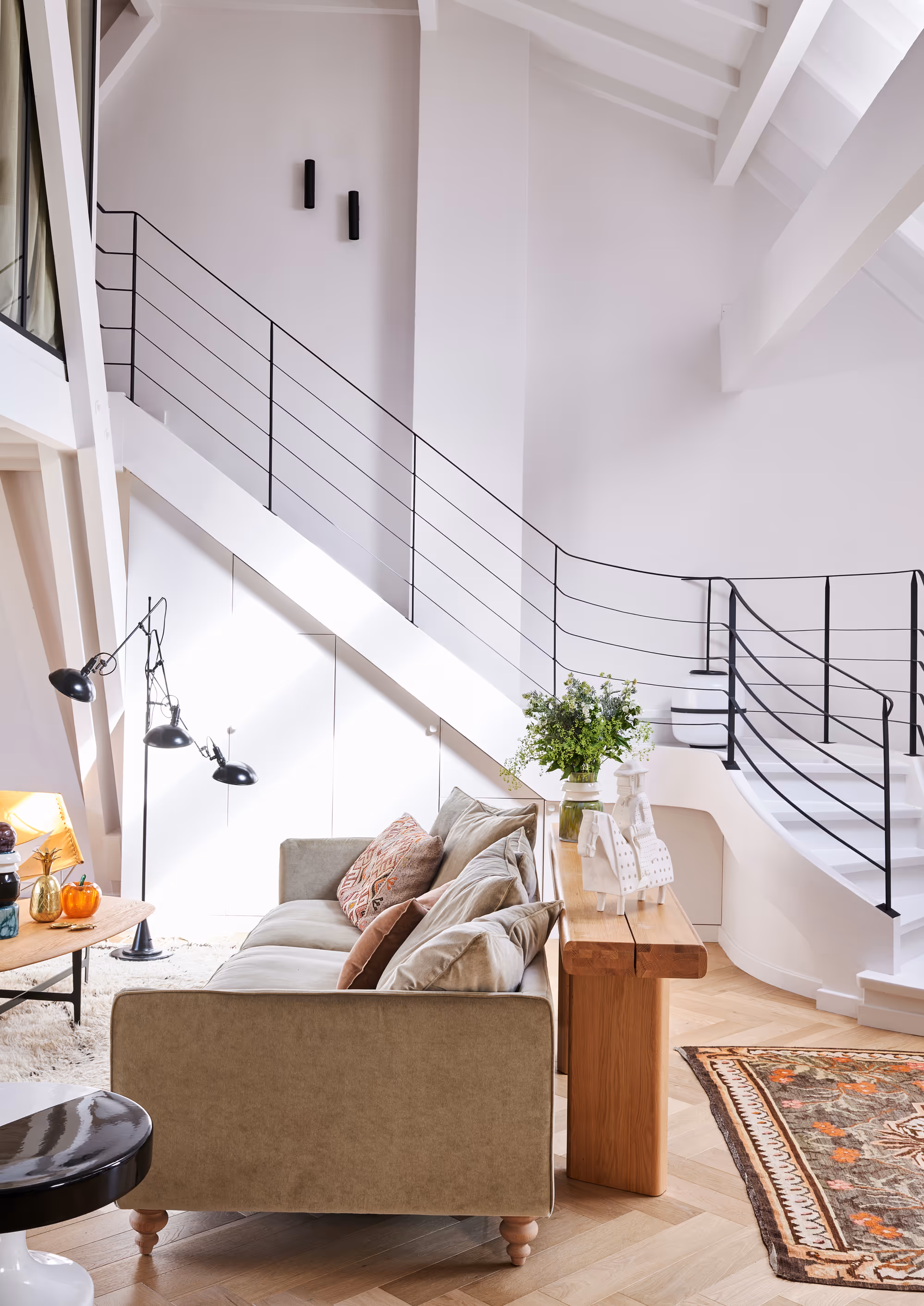 Salon moderne et lumineux d'un duplex à Paris avec canapé beige, tapis à motifs, console en bois avec vases décoratifs et escalier blanc avec rampe noire.