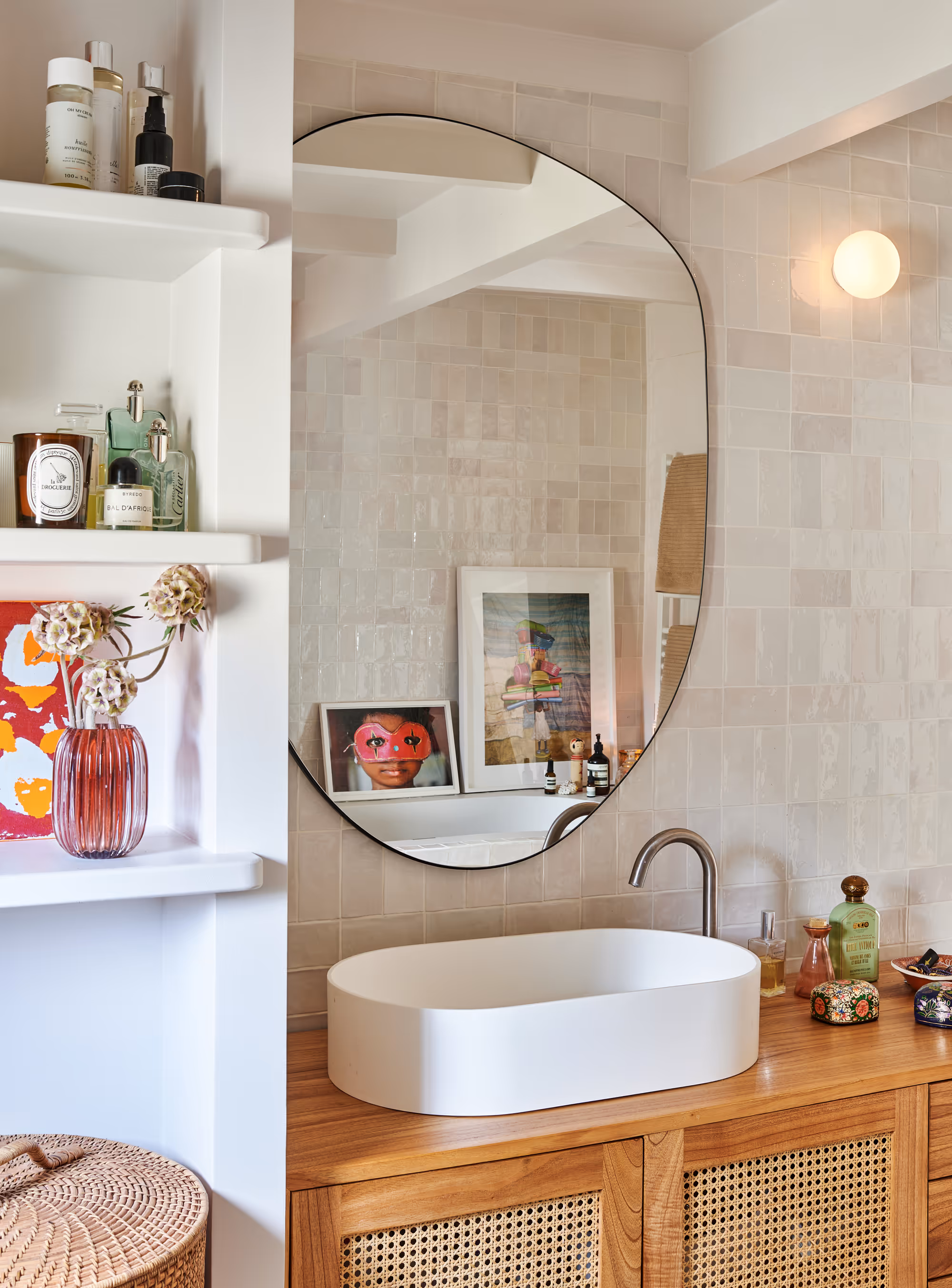 Salle de bain moderne d'un duplex parisien avec meuble-lavabo en bois, lavabo ovale blanc, miroir rond sans cadre et étagères avec des produits de soin pour la peau et des objets décoratifs.