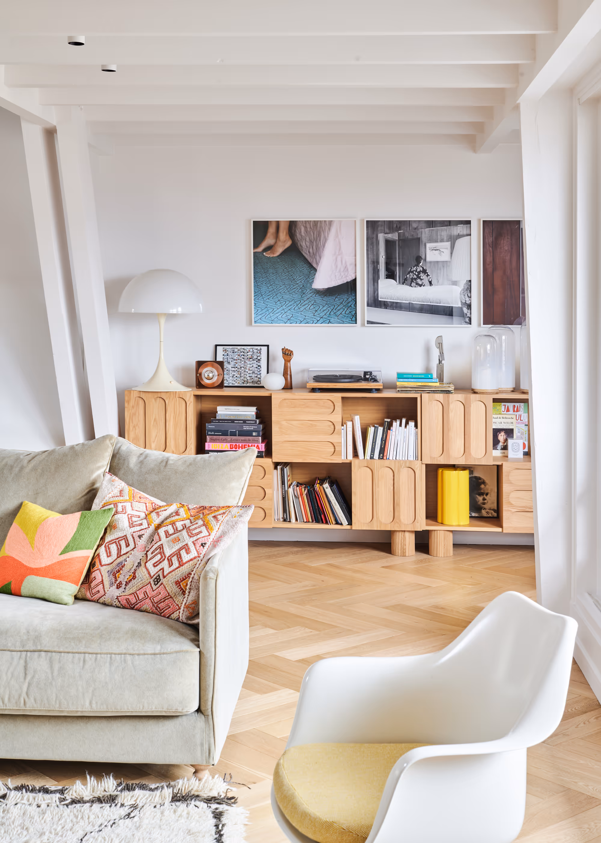 Salon lumineux d'un duplex parisien avec un canapé vert clair orné de coussins colorés, un fauteuil blanc avec un coussin jaune et un meuble en bois sous trois photos encadrées.