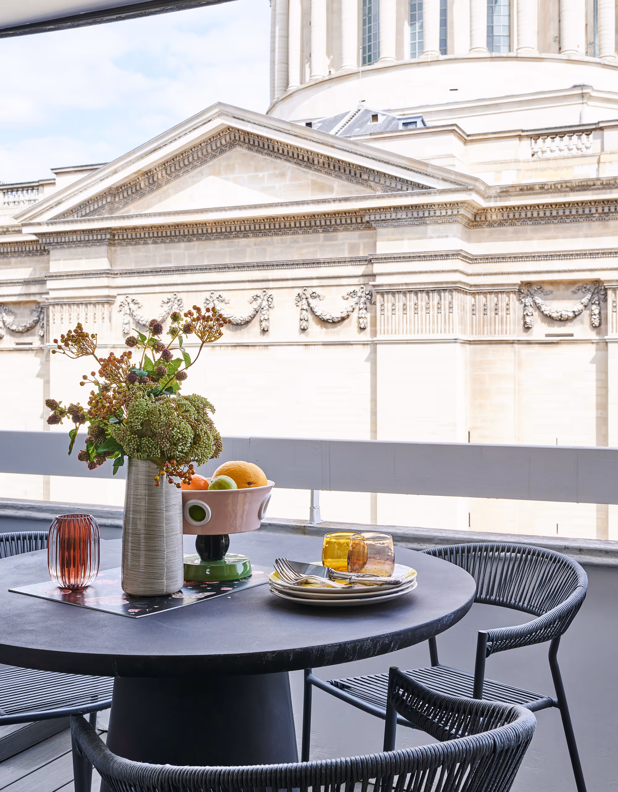 Balcon d'un duplex dans le 5ème arrondissement de Paris. Table ronde noire d'extérieur avec trois chaises, un vase de fleurs, un bol de fruits et des assiettes empilées avec des verres, sur fond de façade architecturale classique.