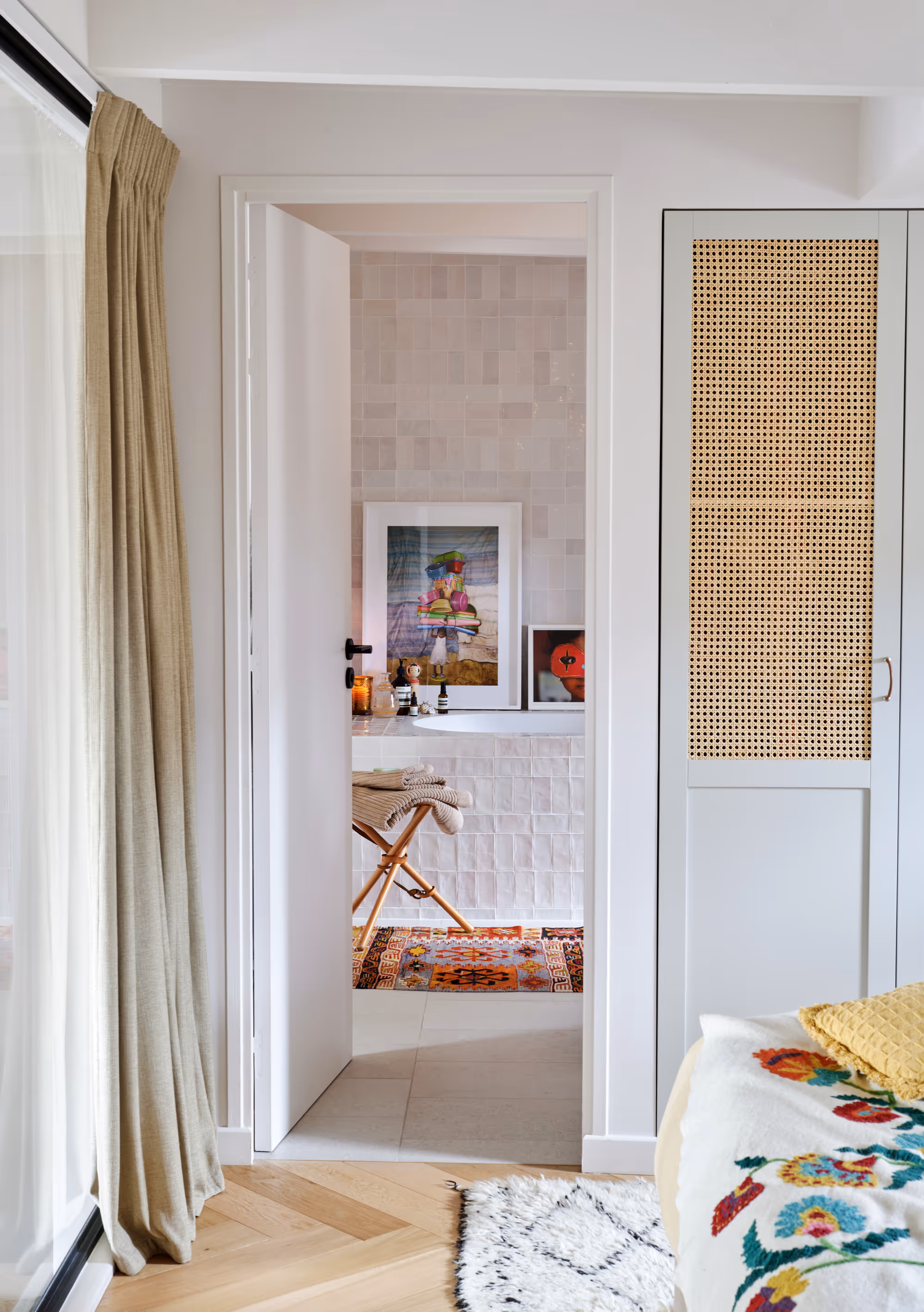 Vue à travers une porte ouverte sur une salle de bain aux murs carrelés de blanc, décorée d'œuvres d'art colorées, avec un tabouret en bois sur lequel sont posées des serviettes pliées et un tapis à motifs, attenante à une chambre à coucher avec une couverture à fleurs et une porte d'armoire en osier.