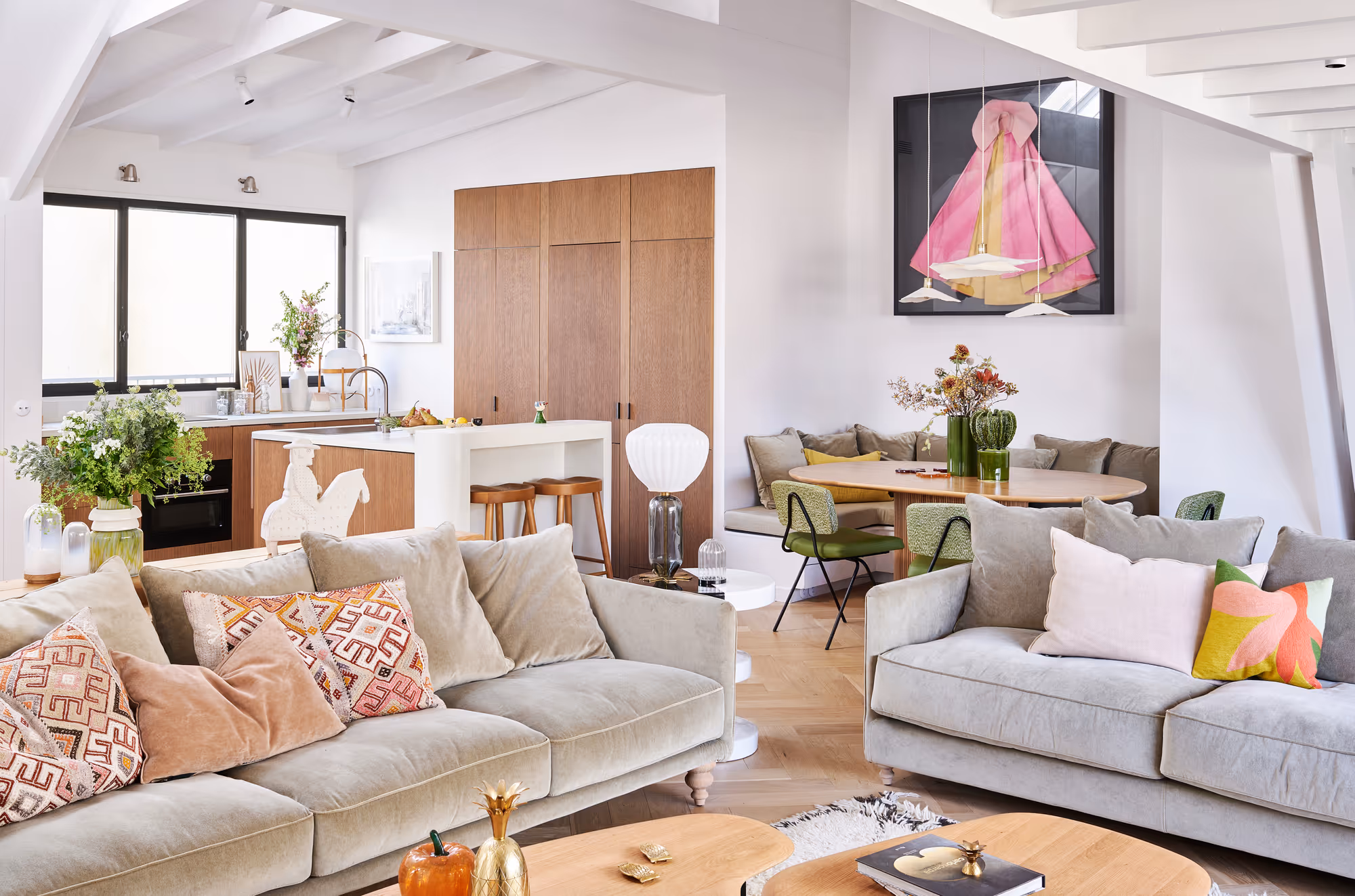 Salon moderne et lumineux d'un duplex parisien avec deux canapés beiges ornés de coussins à motifs, une table basse en bois et un coin repas avec des chaises vertes, à côté d'une cuisine équipée d'armoires en bois et d'un îlot blanc.