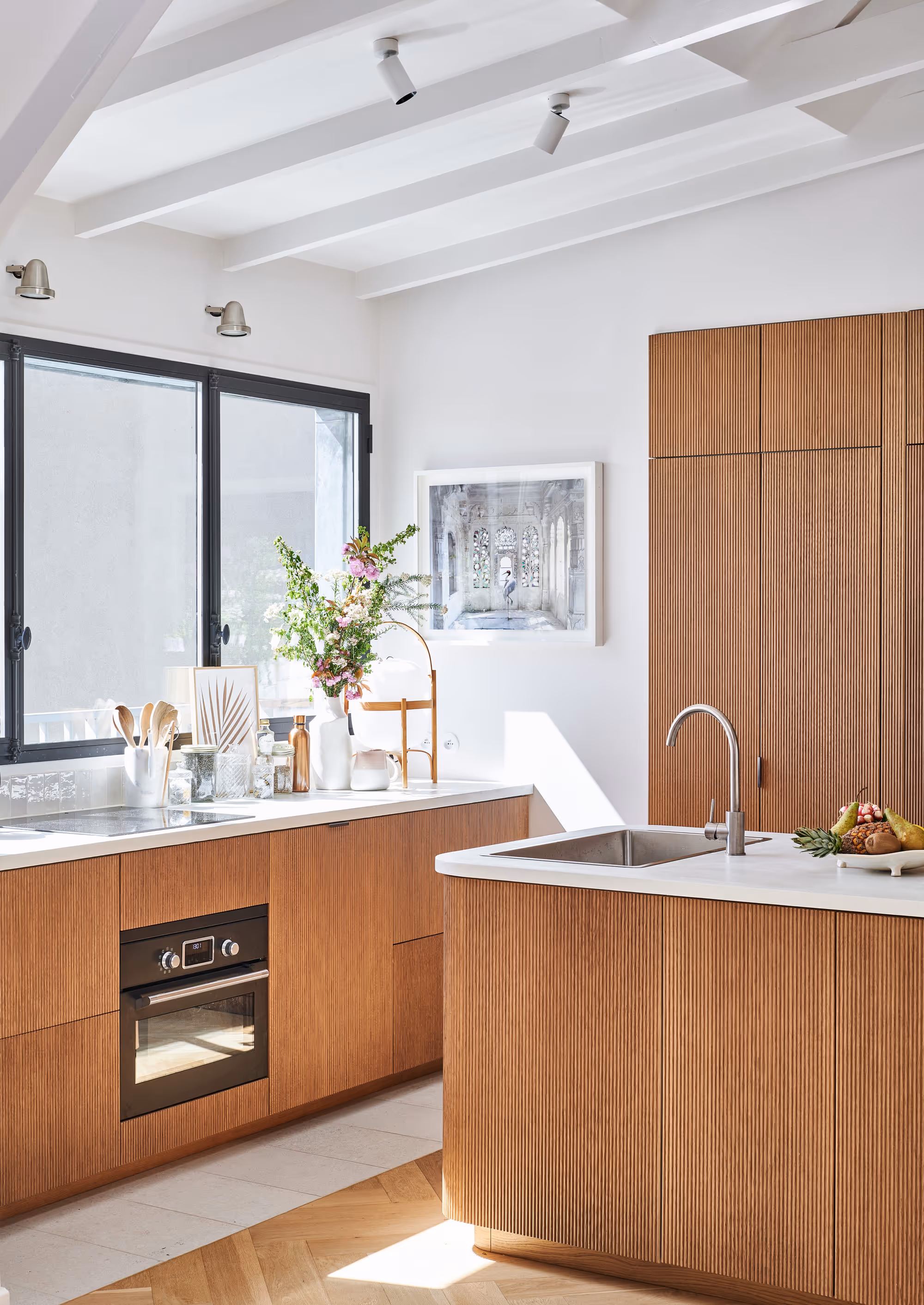 Cuisine moderne et lumineuse d'un duplex à Paris avec des armoires en bois, des plans de travail blancs, un évier en acier inoxydable, un four et un vase de fleurs sur le comptoir sous de grandes fenêtres.