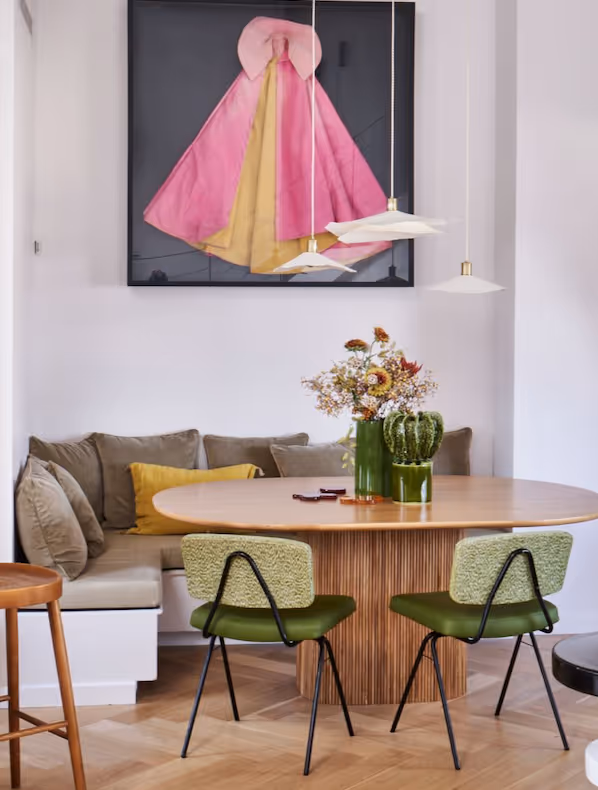Salle à manger moderne d'un duplex à Paris avec une table ronde en bois, des chaises rembourrées vertes, des banquettes encastrées et une œuvre d'art encadrée représentant une cape rose et jaune accrochée à un mur blanc.