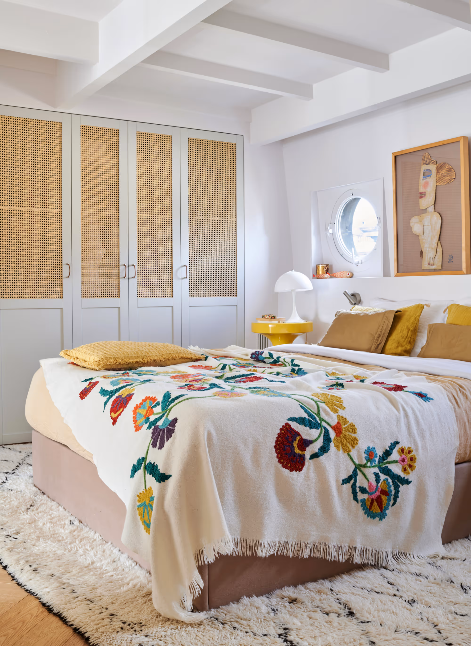 Chambre confortable d'un duplex à Paris avec un lit beige recouvert d'une couverture blanche brodée de fleurs colorées, des coussins moutarde et marron, une table de chevet jaune avec une lampe blanche, une fenêtre ronde et des armoires gris clair avec des panneaux en canne tressée.