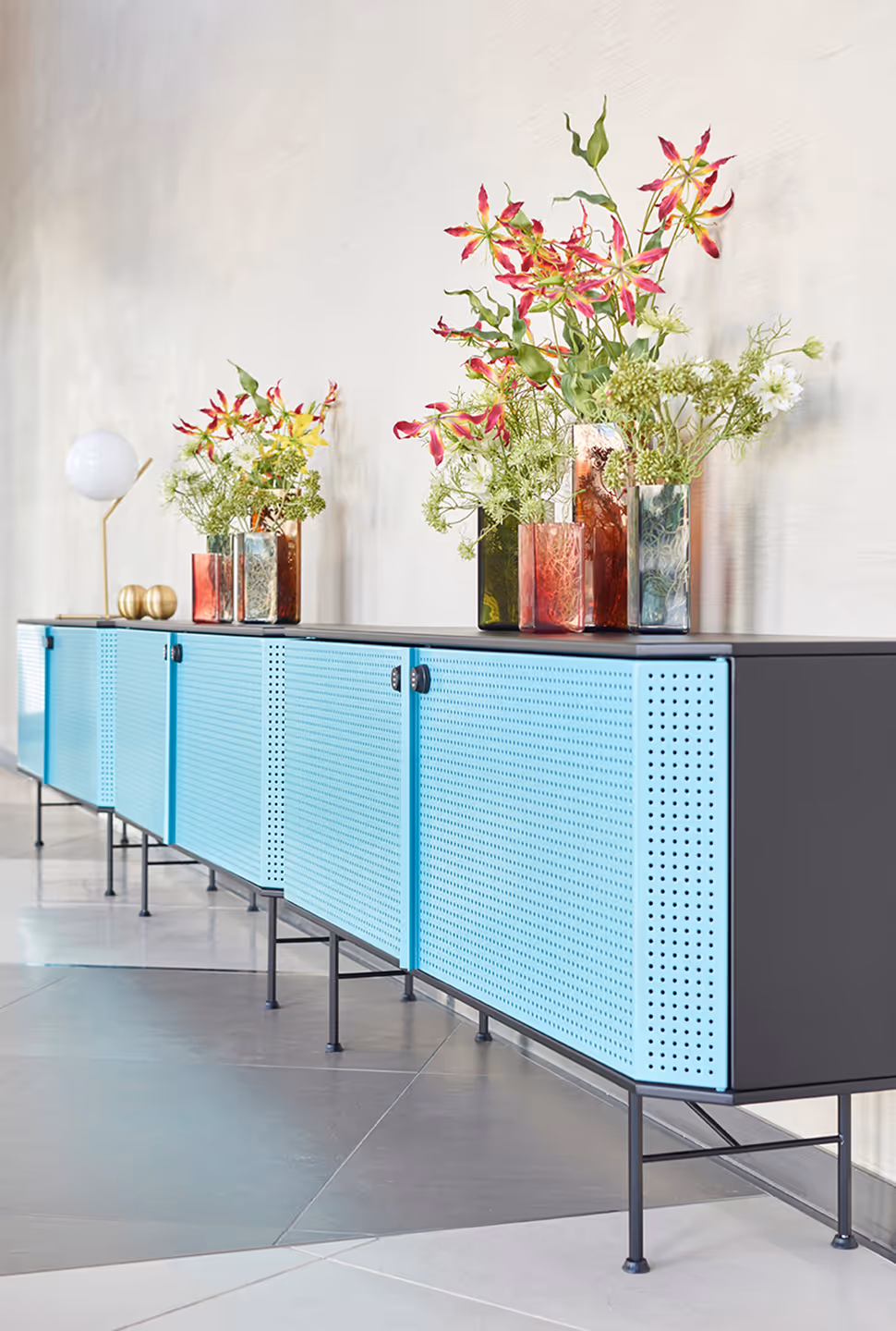 Buffet moderne avec portes perforées bleu clair, plateau noir, orné de vases en verre colorés contenant des fleurs roses et blanches, et une lampe globe blanche à gauche.