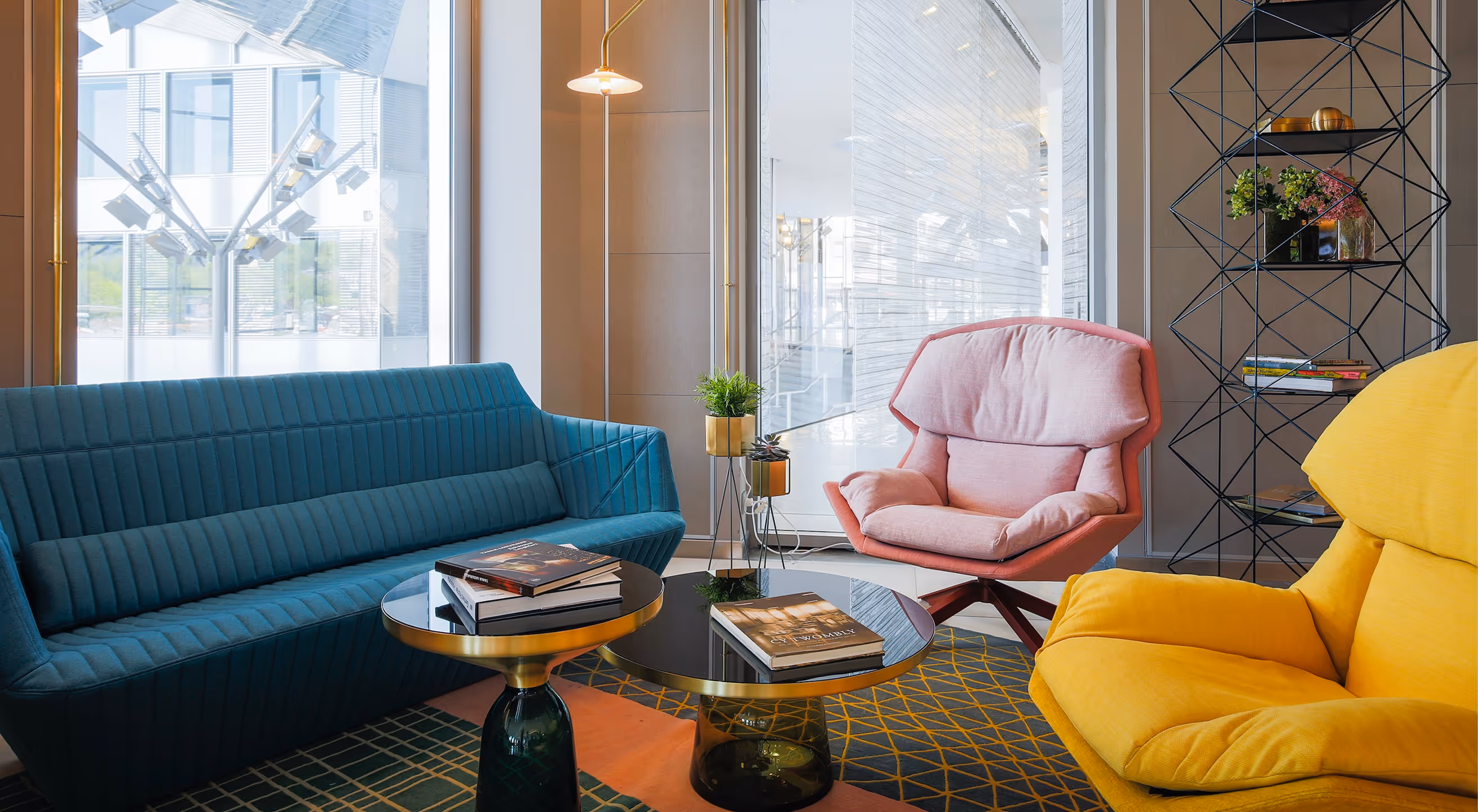 Salon moderne avec un canapé bleu sarcelle, un fauteuil rose, un fauteuil jaune, deux tables basses rondes avec des livres et un grand lampadaire près des grandes fenêtres.