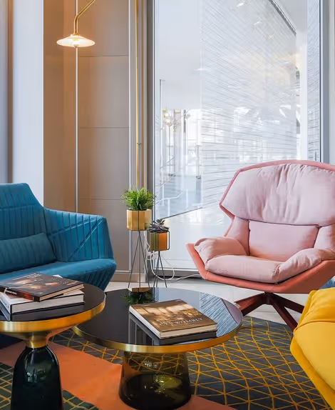 Salon moderne avec un fauteuil bleu sarcelle, une chaise rembourrée rose, un siège jaune, deux tables basses rondes noires et dorées avec des livres et une petite plante en pot sur l'une des tables.