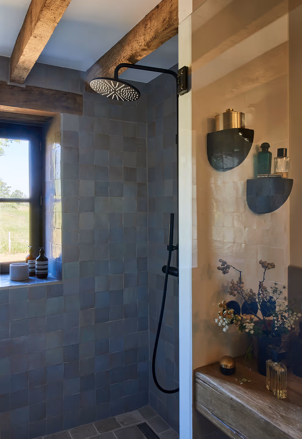Salle de bain rustique avec pomme de douche à effet pluie noir mat, carrelage gris carré, poutres apparentes et fenêtre avec des bouteilles sur le rebord.