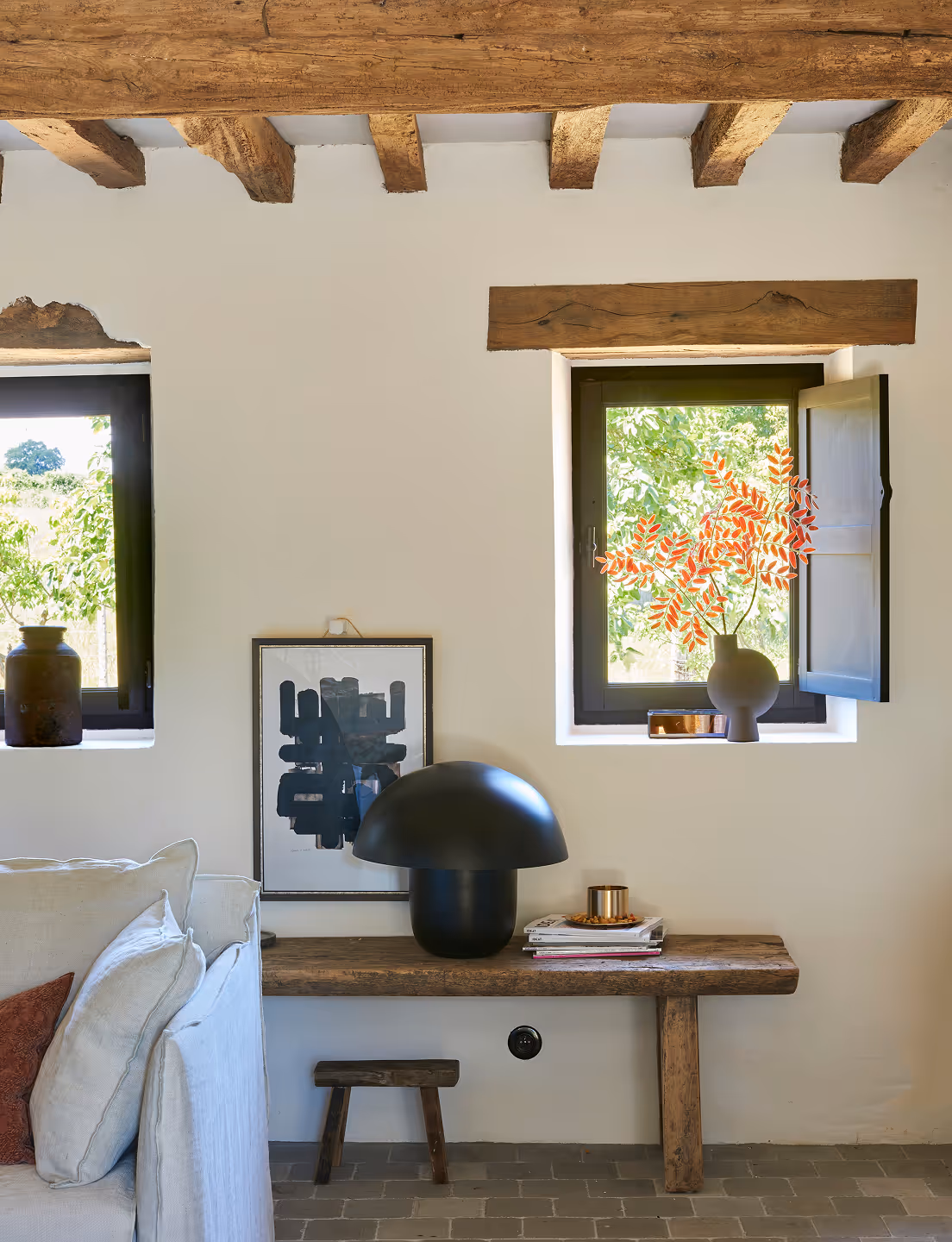 Coin rustique avec plafond à poutres apparentes, deux fenêtres, un banc en bois sur lequel sont posés une lampe noire en forme de champignon, des livres empilés et un vase contenant des feuilles orange, à côté d'un canapé beige avec des coussins.