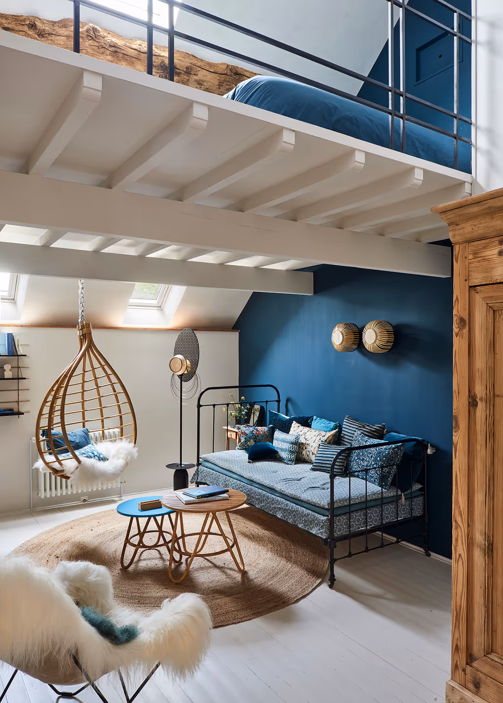 Salon confortable avec une chaise suspendue en osier, un canapé à structure métallique avec des coussins bleus, des tables d'appoint en bois et un fauteuil blanc moelleux sous un lit mezzanine.