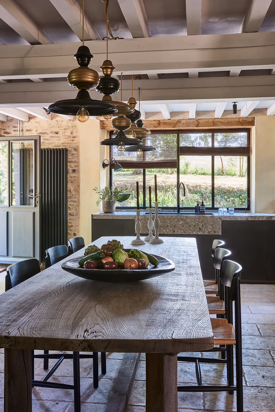 Coin cuisine rustique avec une grande table en bois, des chaises noires, des suspensions lumineuses et une fenêtre donnant sur la verdure.