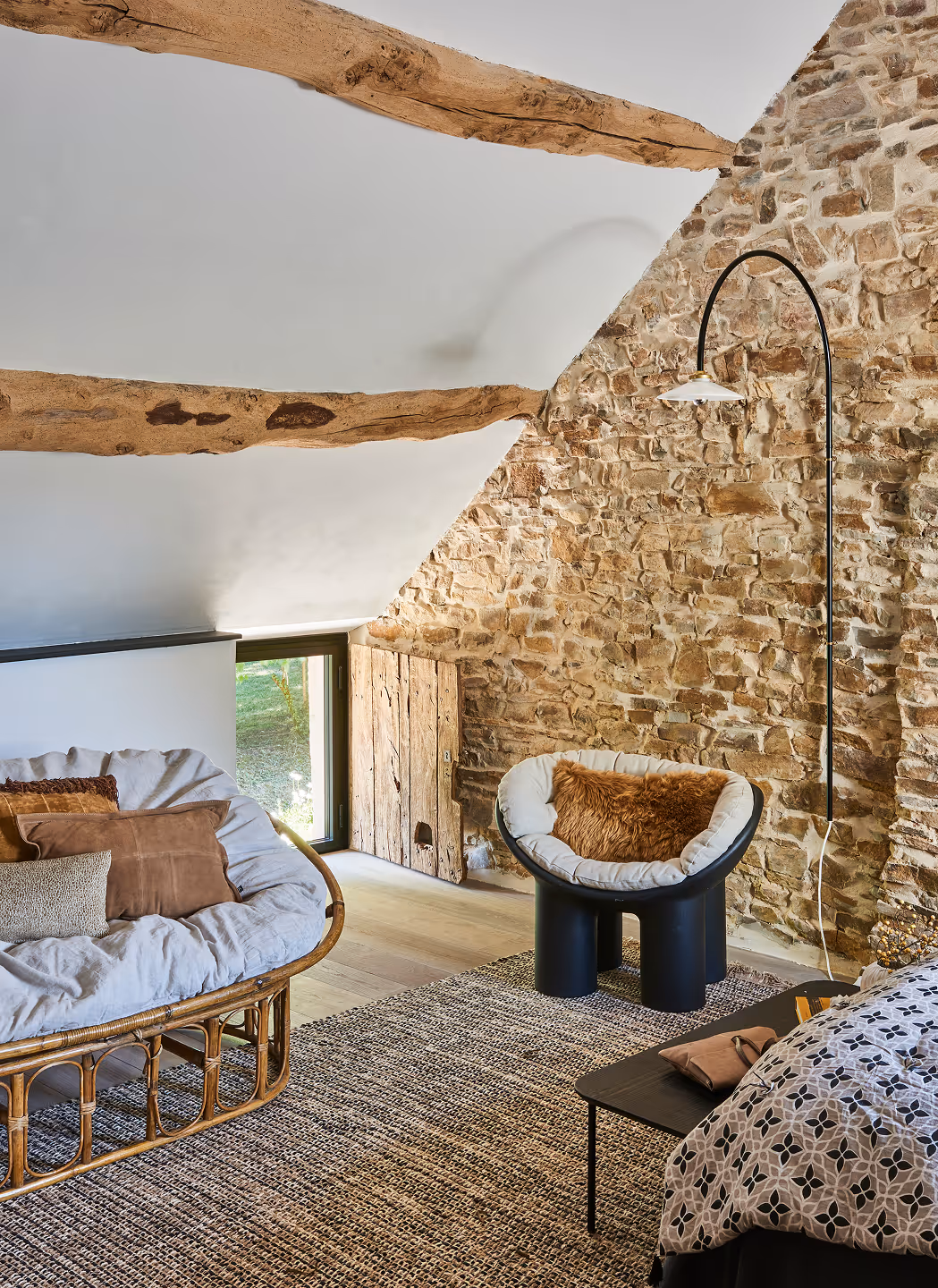 Chambre mansardée confortable avec mur en pierres apparentes, poutres en bois, canapé en rotin rembourré, fauteuil noir moderne avec un coussin marron et couvre-lit à motifs.