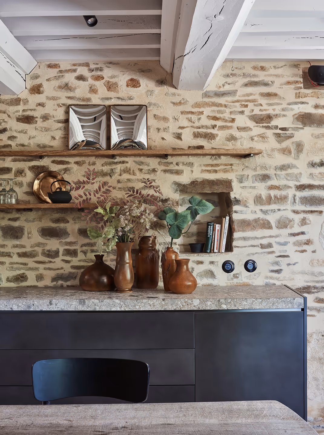 Intérieur de cuisine rustique avec mur en pierre, étagères en bois supportant des vases, une théière, de la verrerie et des livres, et une chaise noire devant un comptoir en pierre.