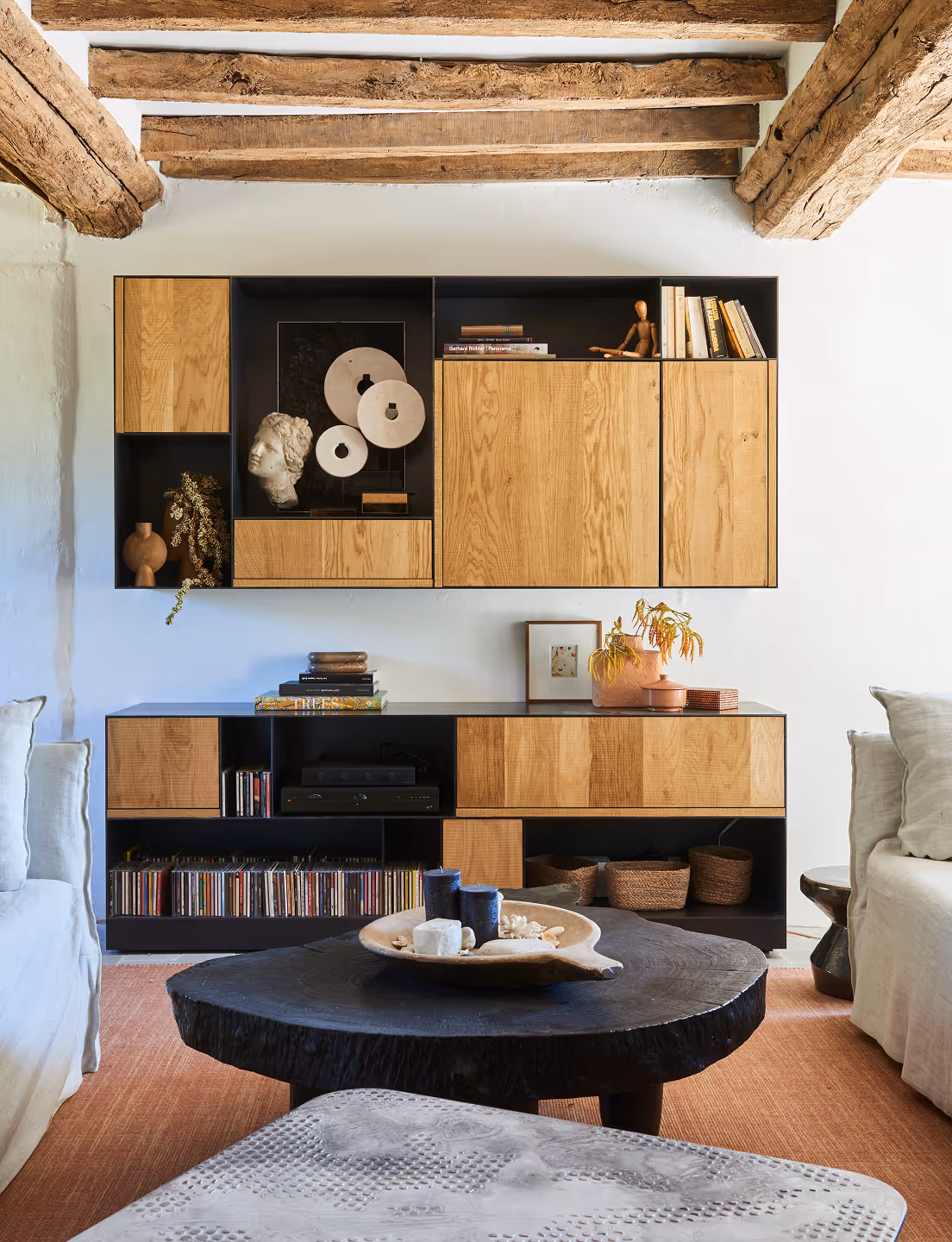 Salon avec étagères flottantes en bois, sculptures décoratives, livres et table basse ronde en bois avec bougies et coquillages.
