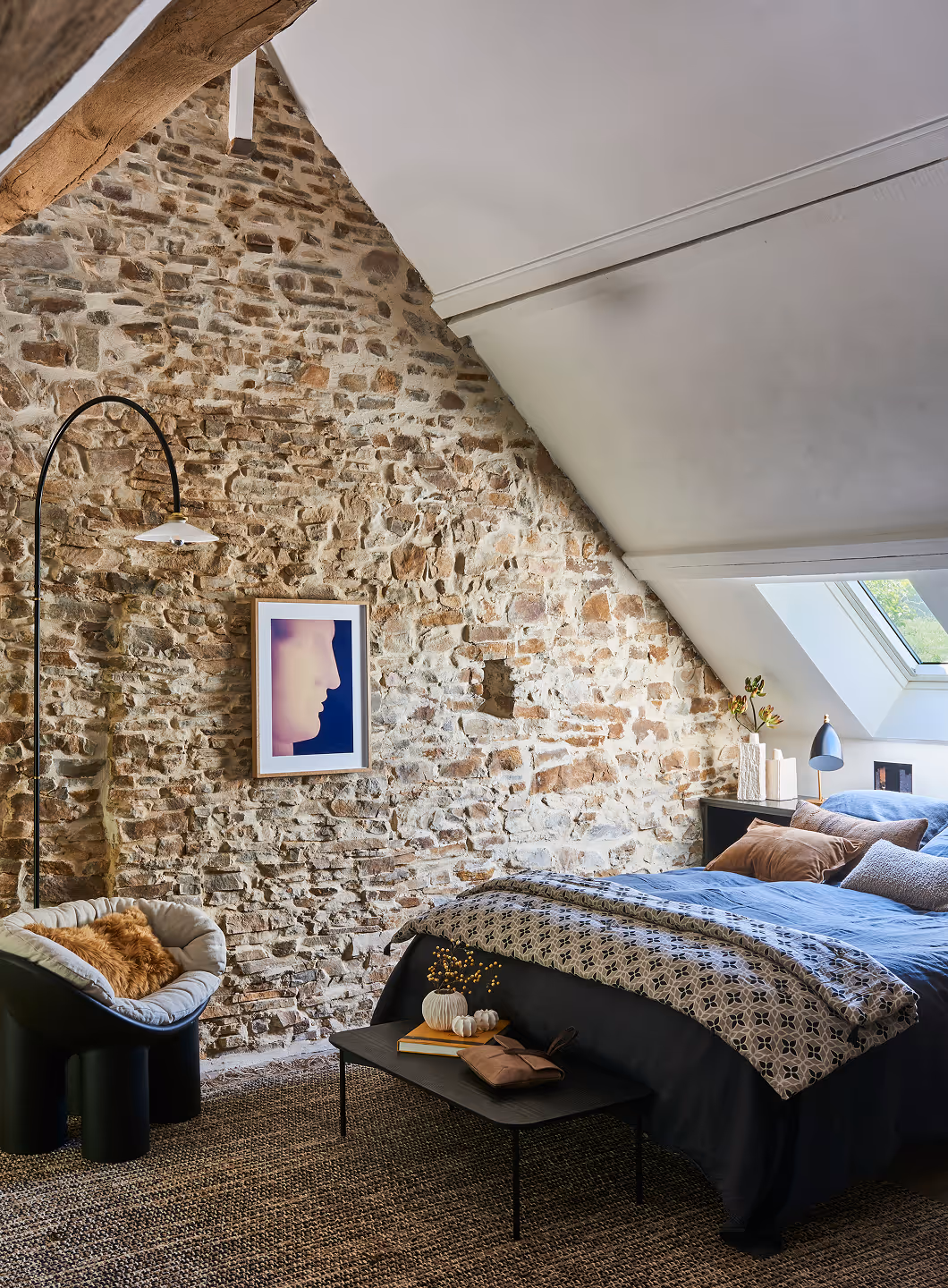 Chambre confortable avec un plafond en pente, un mur en pierres apparentes, une literie bleue, des coussins décoratifs et une chaise noire recouverte d'un plaid rembourré.