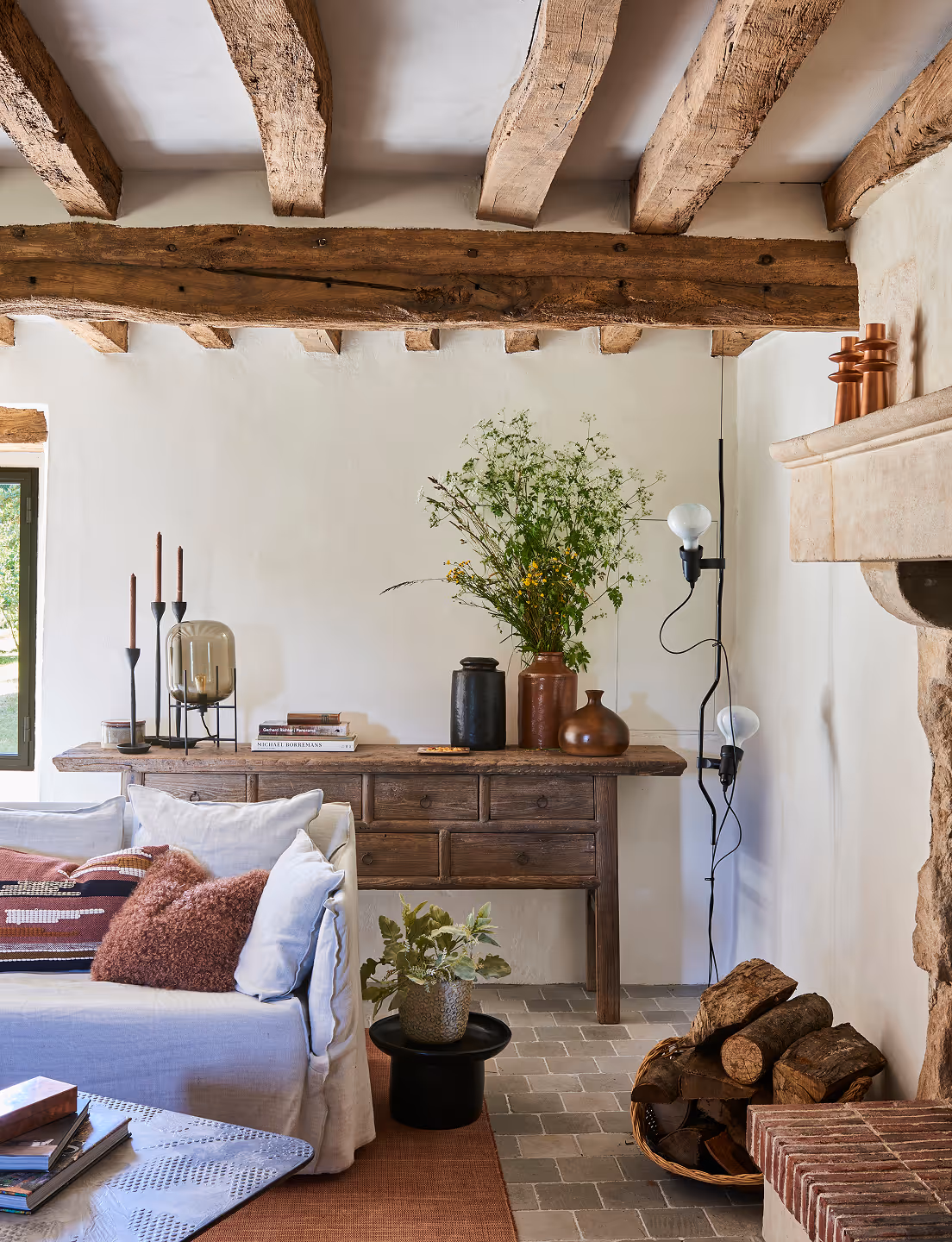 Coin salon confortable avec canapé blanc, console en bois avec vases et bougies, ampoules suspendues, poutres apparentes et panier de bois près d'une cheminée en pierre.