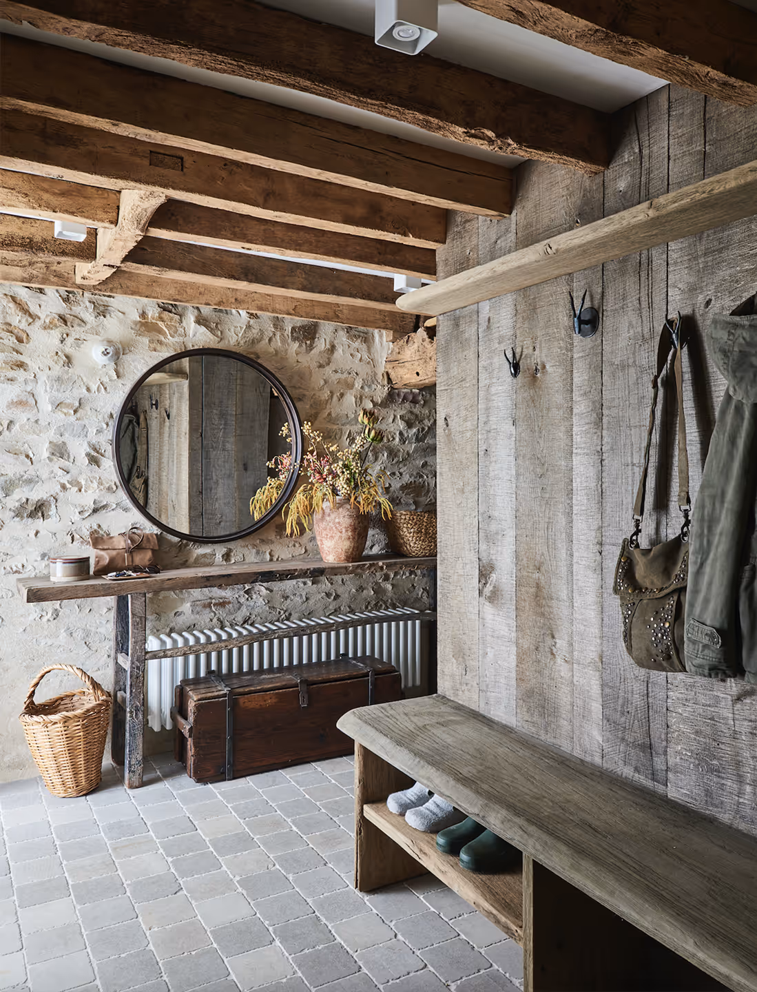 Entrée rustique avec banc en bois, murs en pierre, miroir rond, panier en osier et coffre en bois sous la table.