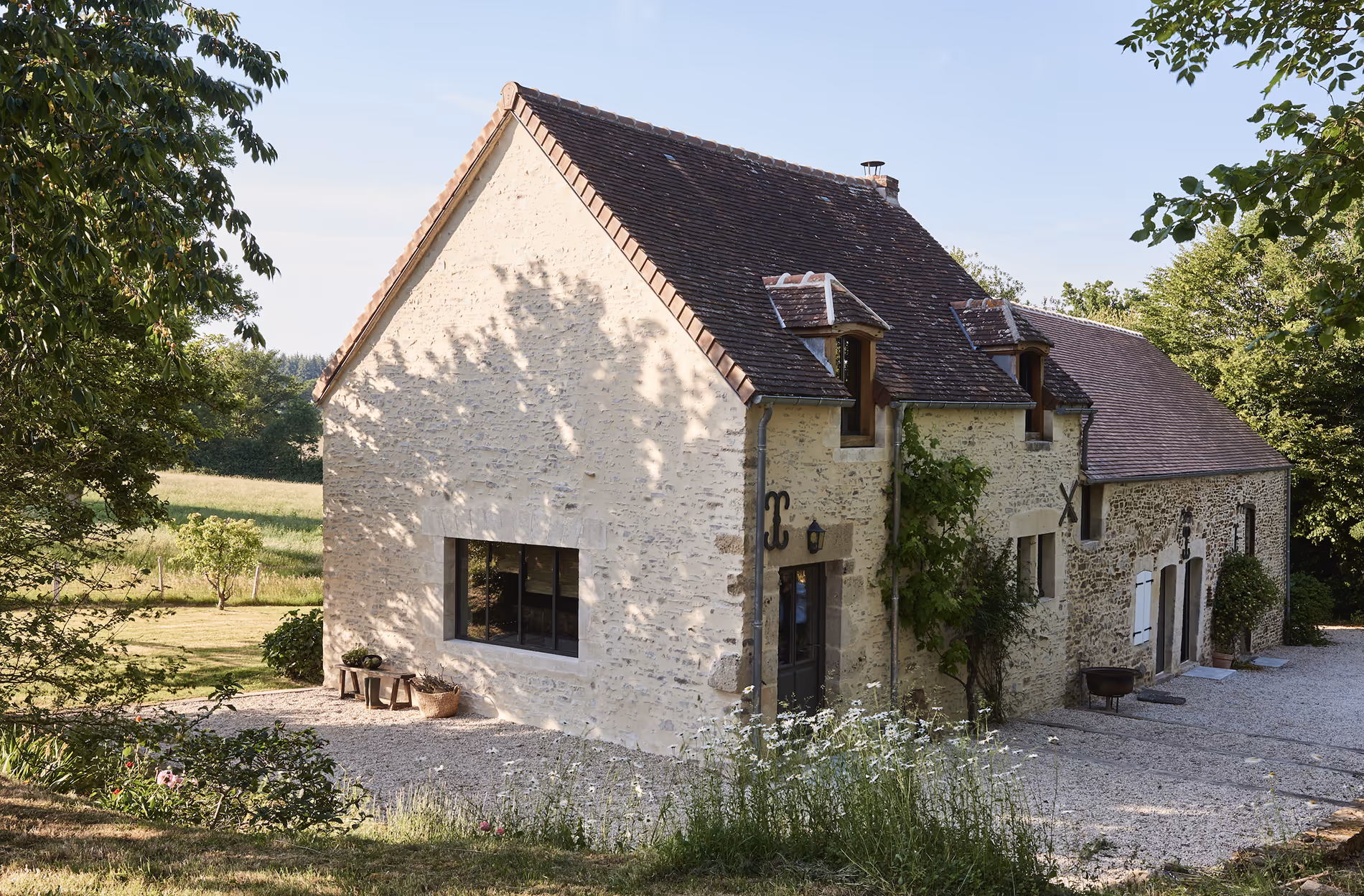 Maison de campagne en Normandie en pierre avec un toit de tuiles sombres, entourée de verdure et d'une allée de gravier, à la lumière du jour.