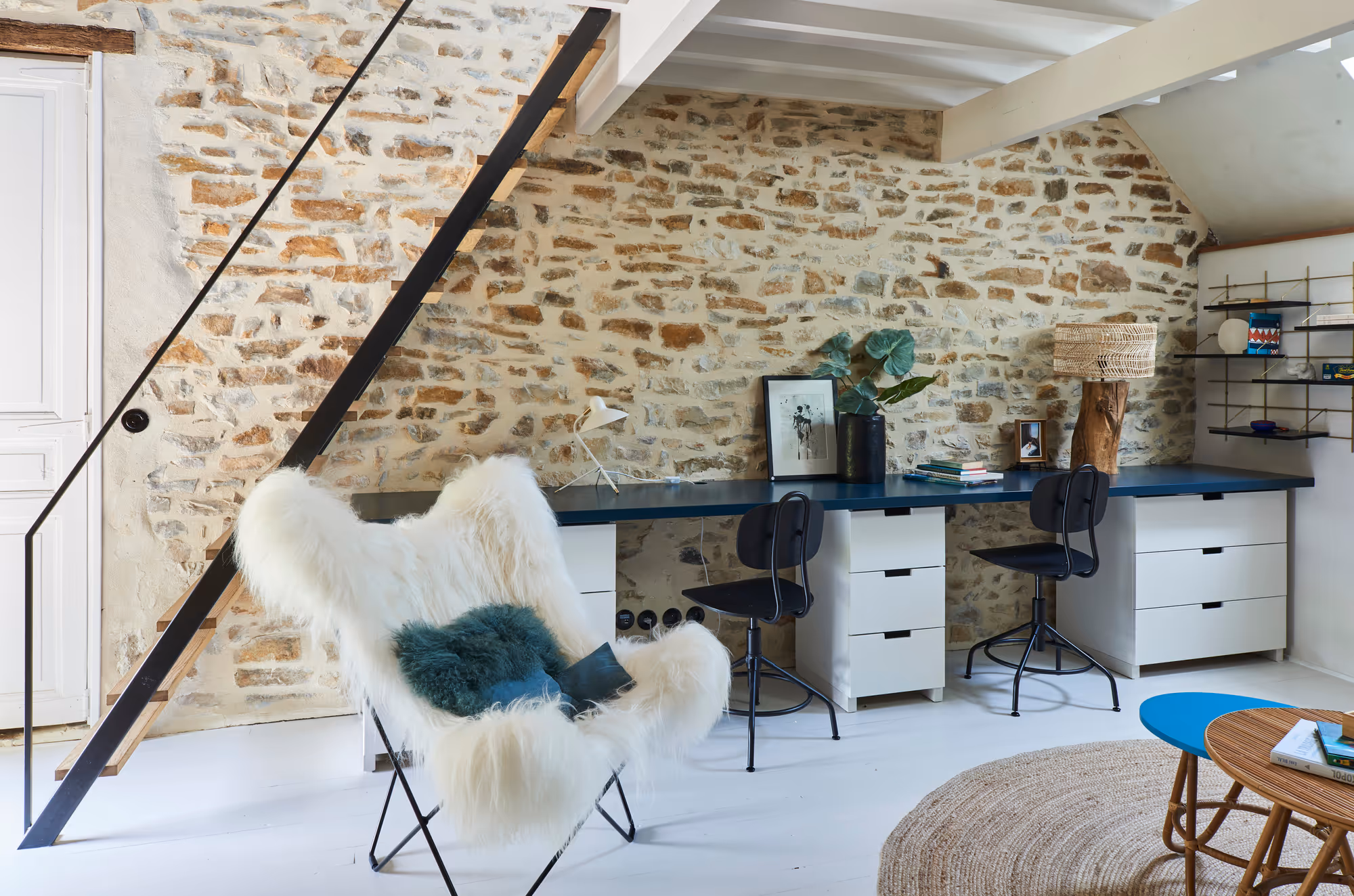 Bureau à domicile rustique et confortable avec un mur en pierre, un long bureau bleu avec deux chaises noires, une chaise papillon en fourrure et un tapis circulaire tissé.