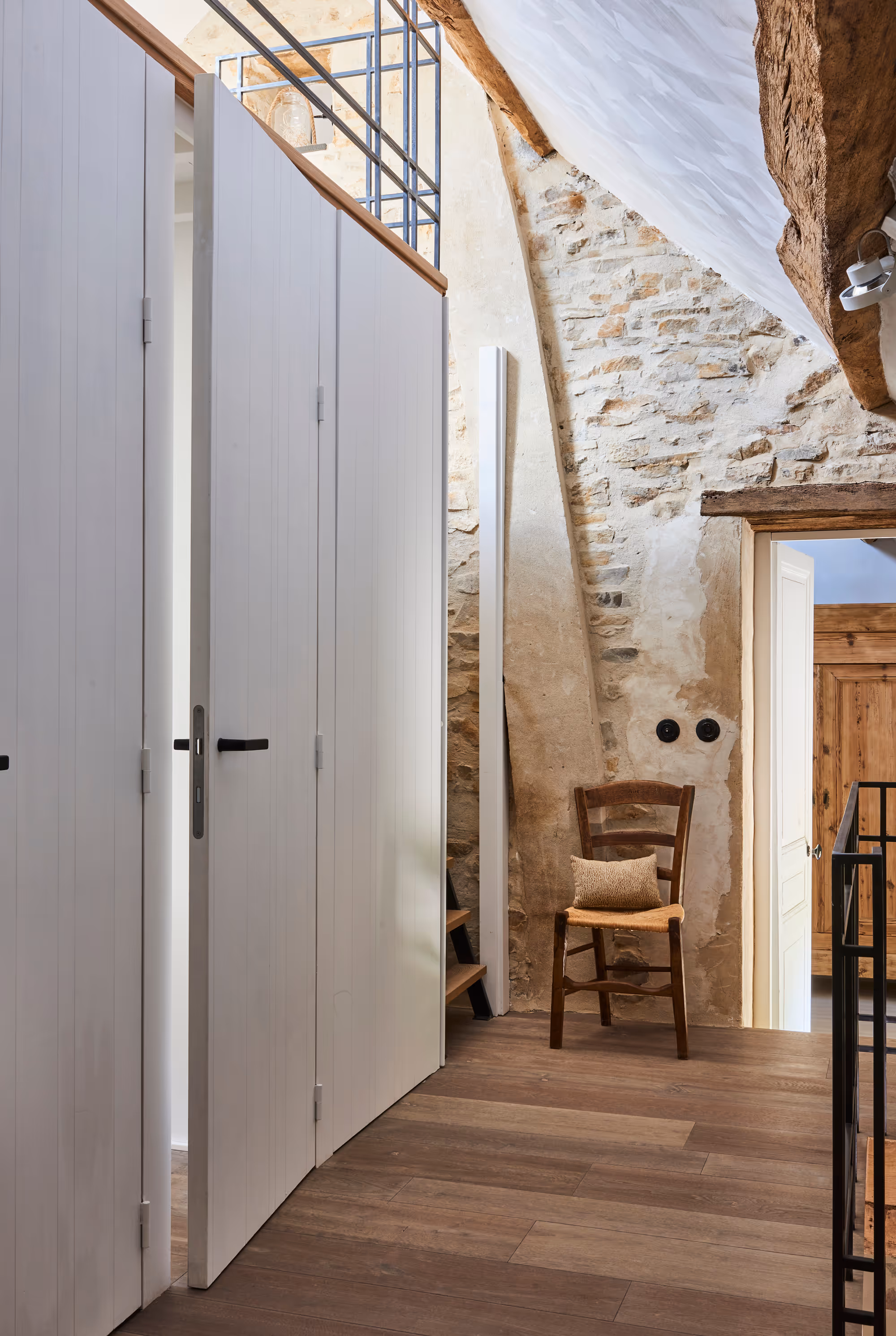Couloir intérieur avec murs en pierre rustiques, sol en bois, portes à panneaux blancs et chaise en bois avec coussin.