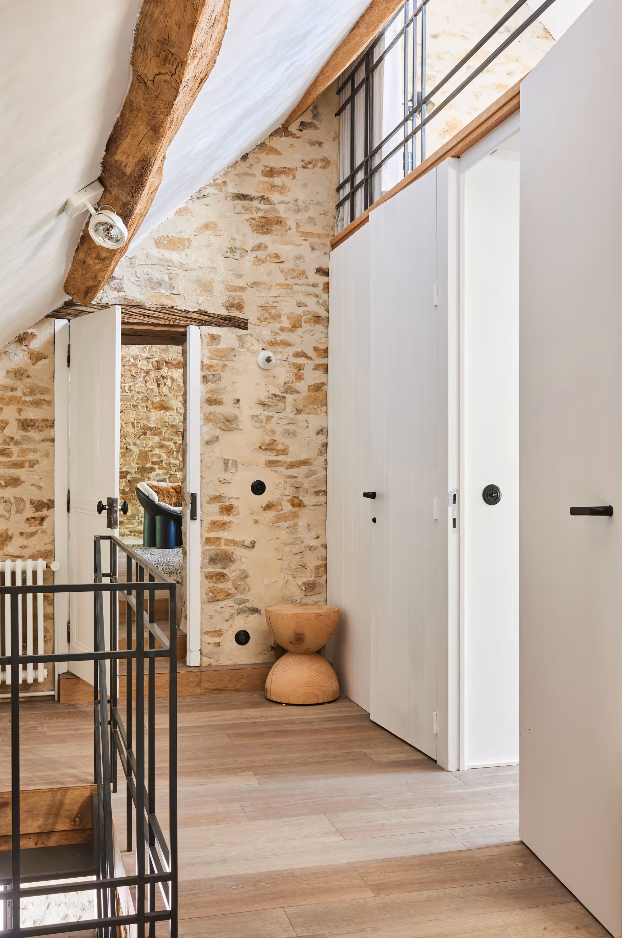 Couloir lumineux avec mur en pierres apparentes, poutres en bois, parquet clair, portes blanches et petit tabouret en bois.