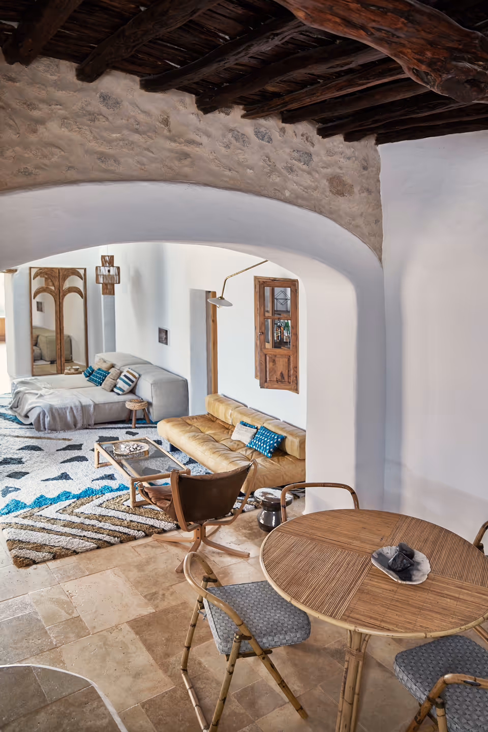 Salon confortable avec poutres apparentes en pierre, canapé en cuir, canapé gris, tapis à motifs et table à manger ronde en bois avec chaises.