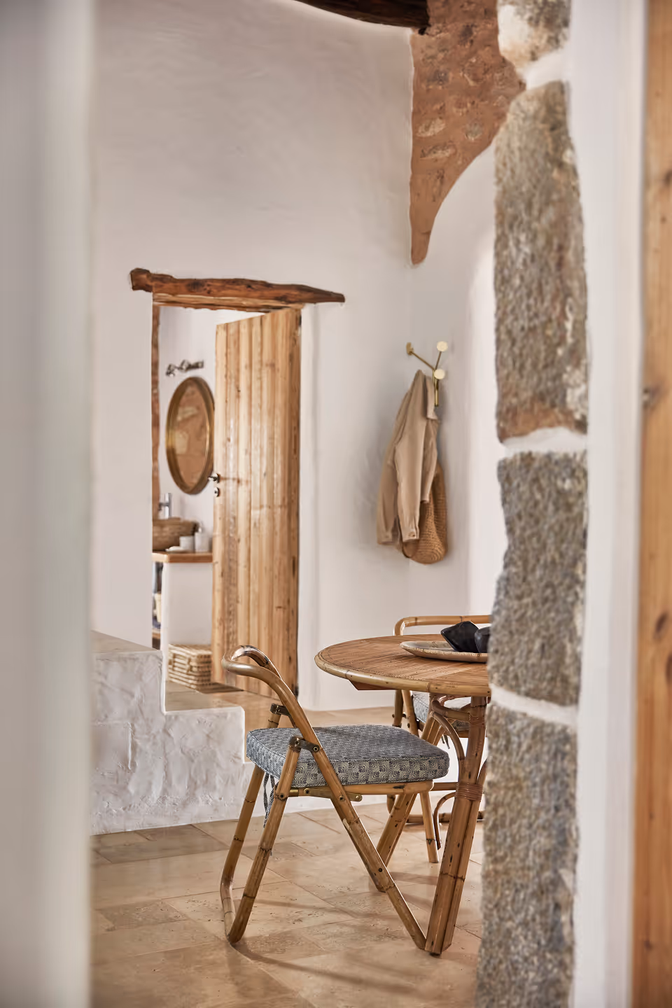 Intérieur rustique avec une table ronde en bois et une chaise pliante rembourrée près d'un mur en pierre et d'une porte en bois ouverte menant à une salle de bain avec un miroir rond.