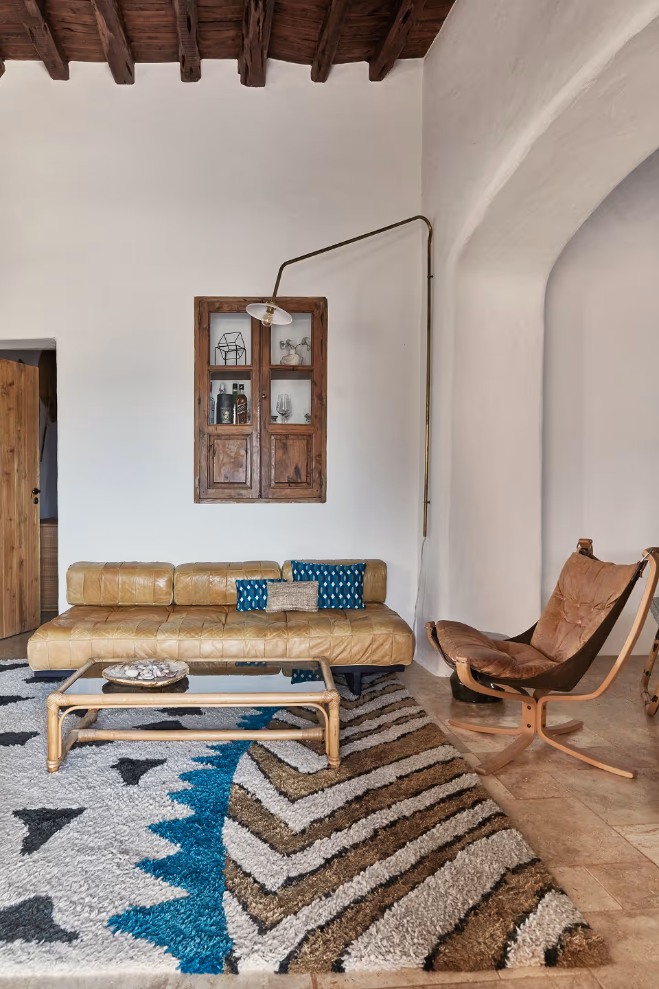 Salon moderne avec un canapé en cuir beige, un tapis à motifs, une table basse en bois et un fauteuil rembourré marron, le tout contre des murs blancs et des poutres apparentes en bois.
