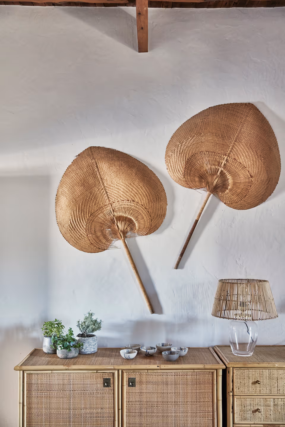 Deux grands éventails tissés accrochés à un mur blanc au-dessus d'un buffet en osier contenant de petits bols, des plantes en pot et un abat-jour en osier sur un socle en verre.