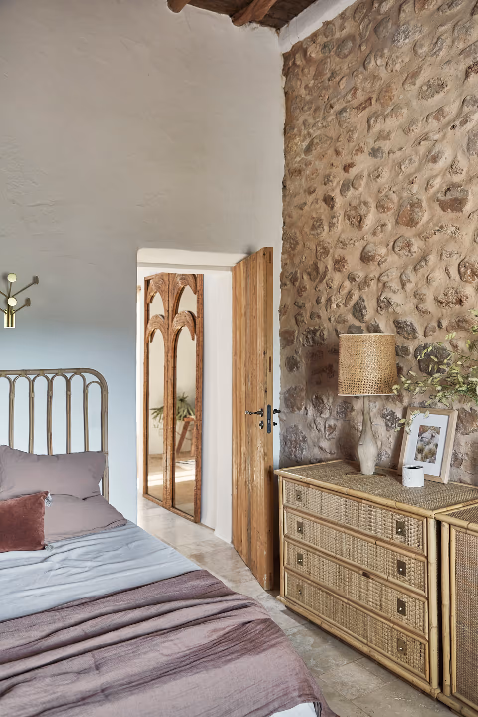 Chambre à coucher avec un cadre de lit en métal, une literie mauve, une commode en bois avec une lampe en rotin, un cadre photo et un mur en pierre.