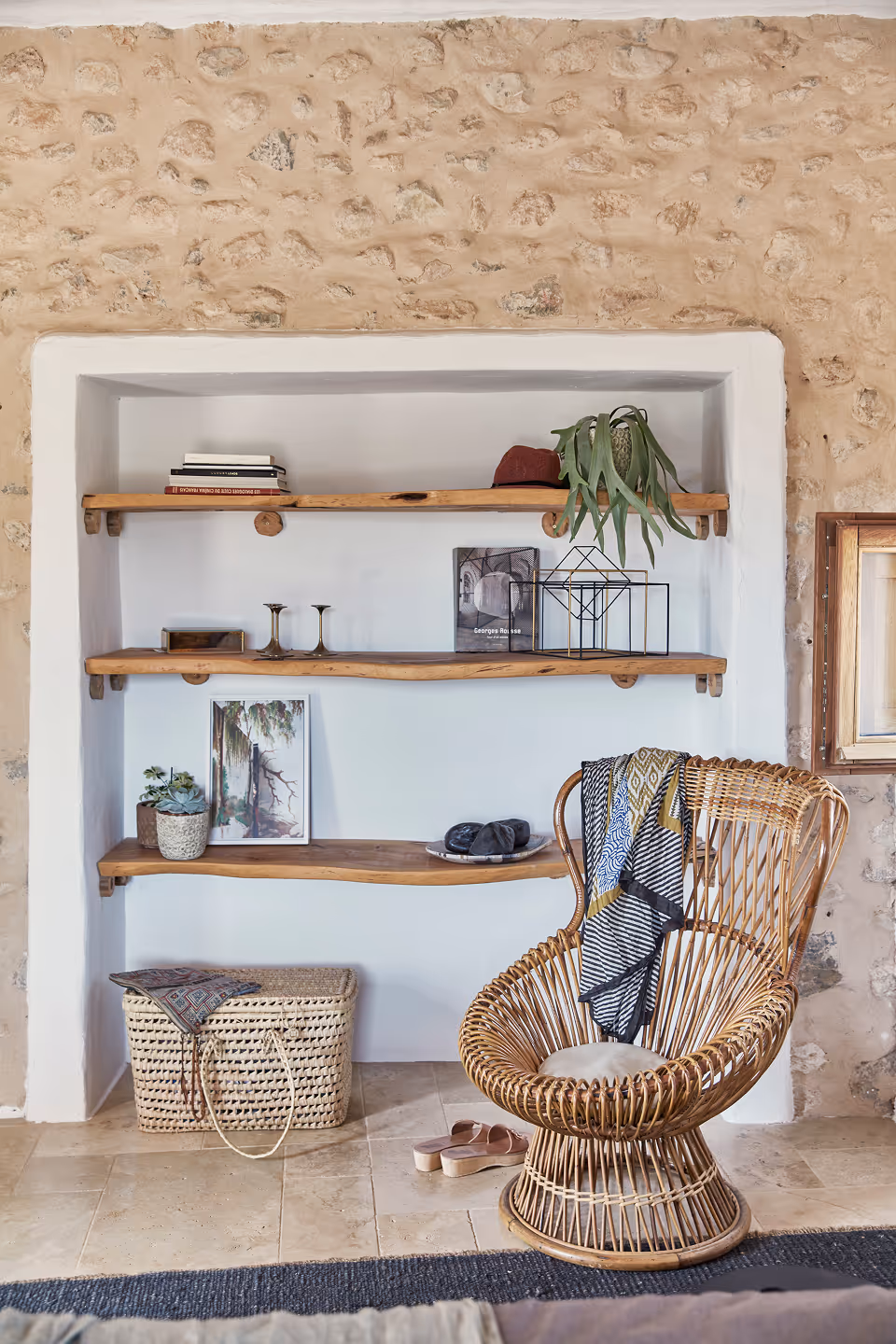 Coin confortable avec une chaise en rotin tressé recouverte d'un foulard à motifs, des étagères murales en bois exposant des livres, des plantes, des bougies et des objets décoratifs, à côté d'un panier tressé posé sur un sol carrelé.
