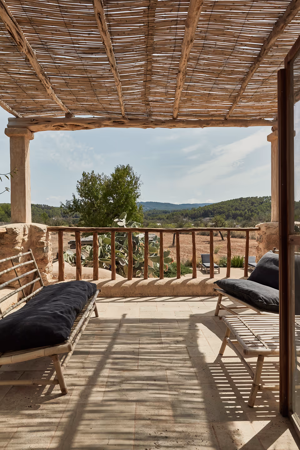 Terrasse rustique ensoleillée avec toit en bambou, bancs en bois avec coussins noirs, donnant sur les arbres et les collines sous un ciel bleu.