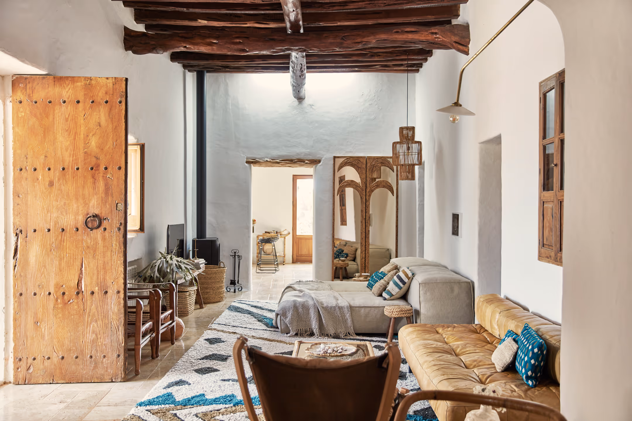 Salon rustique avec poutres apparentes, grande porte en bois, canapé beige, canapé en cuir jaune avec coussins à motifs bleus et tapis texturé.